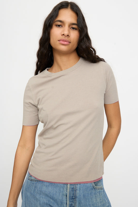 A person with long dark hair wears the B Sides Double Tee in Pink and Dove Grey with blue jeans, standing against a plain light background.