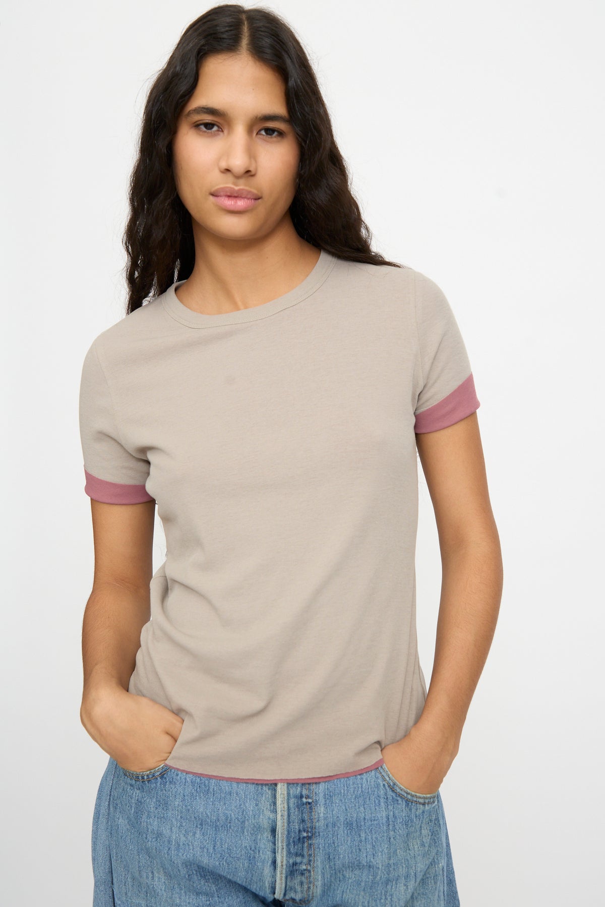 Person with long dark hair wears the Double Tee in Pink and Dove Grey by B Sides and blue jeans, hands in pockets, against a plain light background.