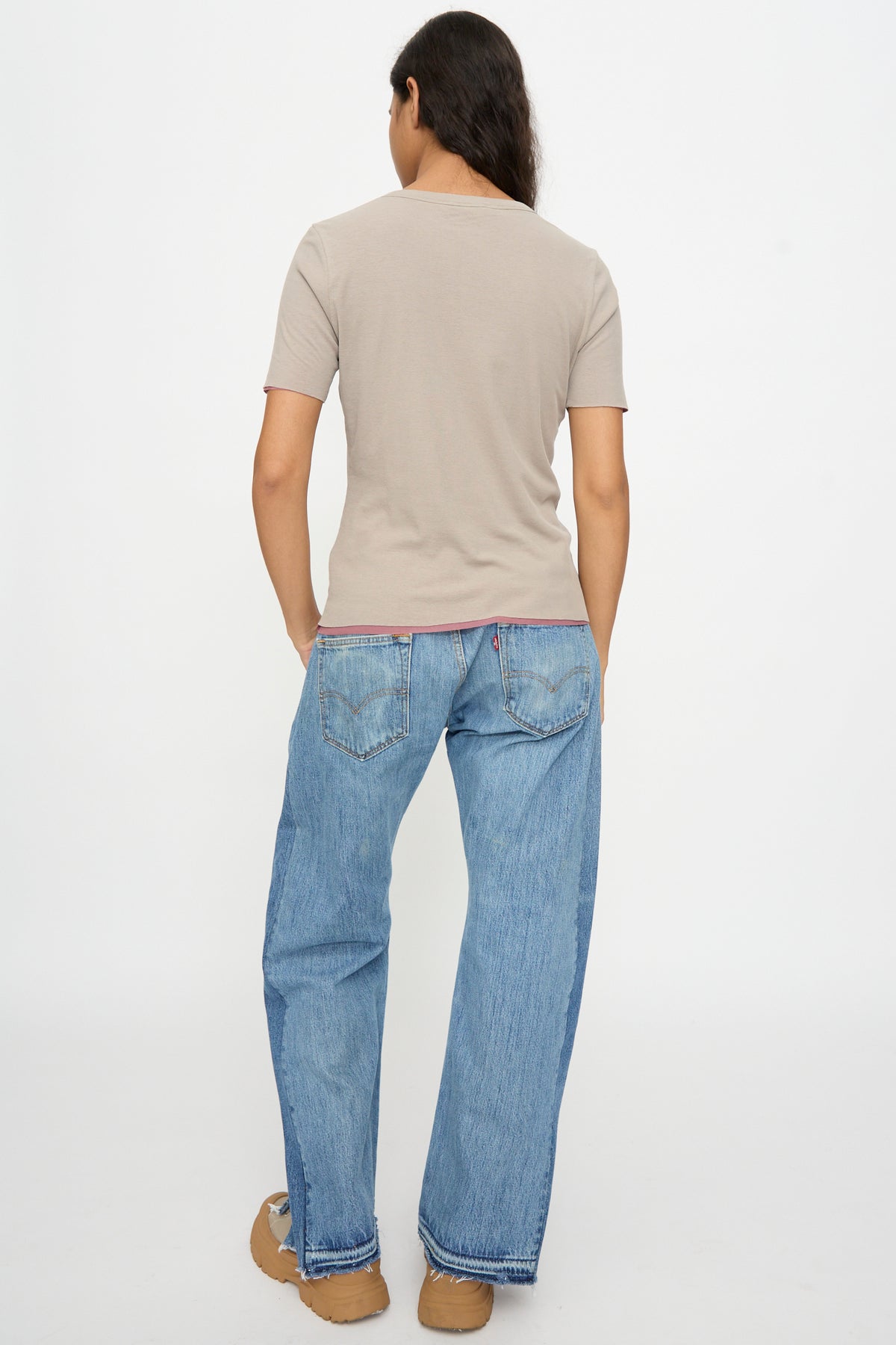 Person stands with their back to the camera, wearing the B Sides Double Tee in Pink and Dove Grey, blue jeans, and tan shoes against a plain white background.