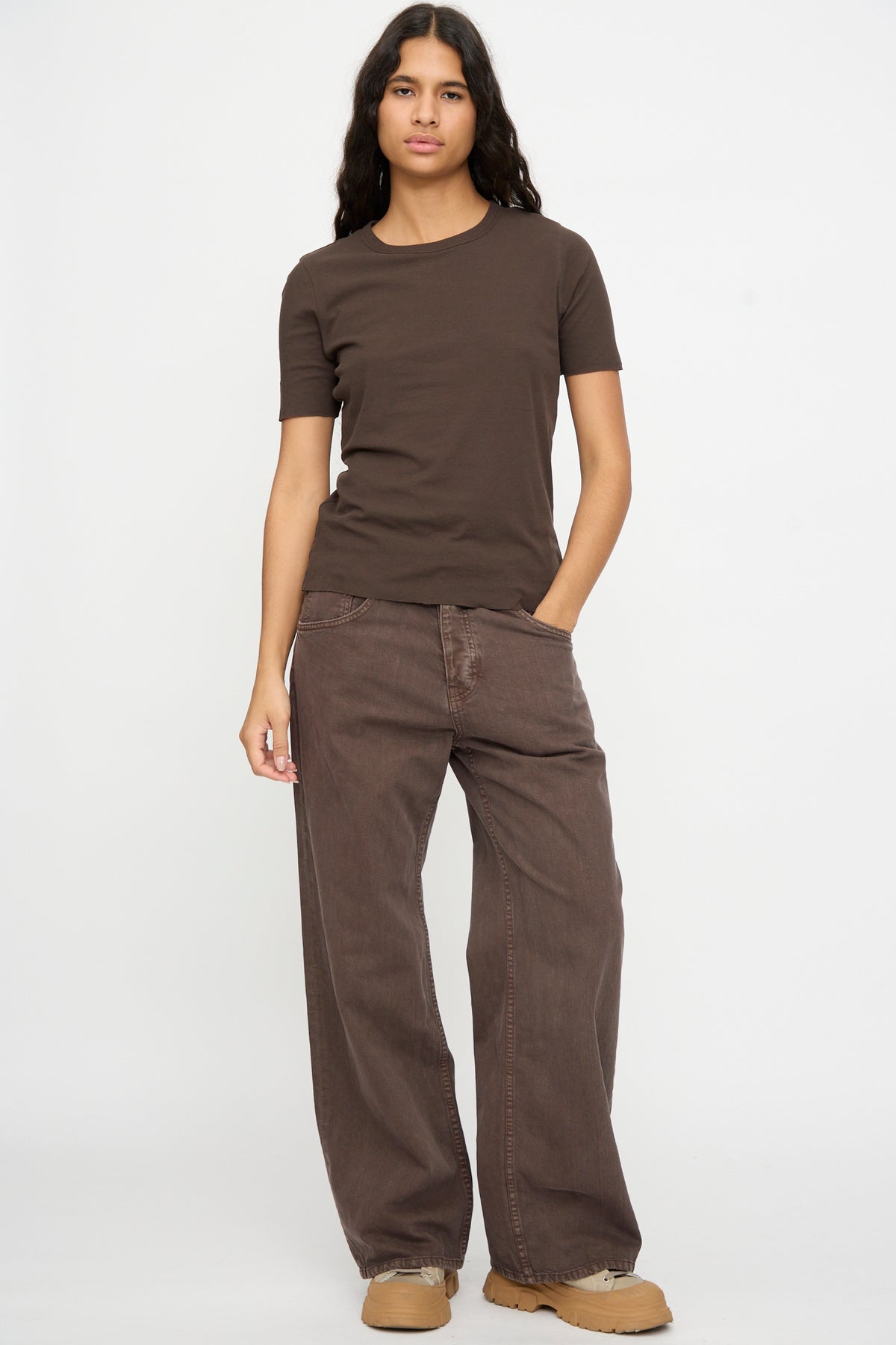 A person stands against a white background wearing the B Sides Double Tee in Walnut and Black, loose brown pants, tan shoes, and has one hand in their pocket.