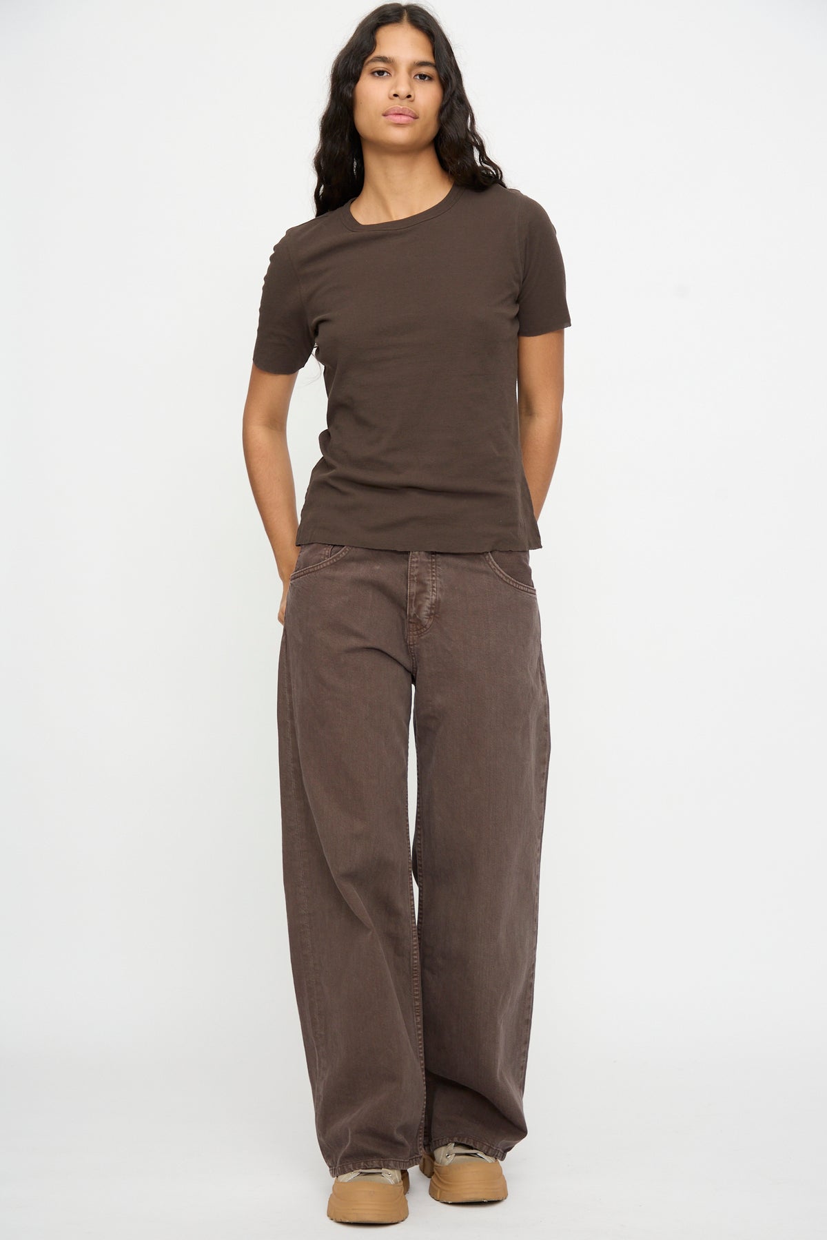 Person stands against a plain background wearing B Sides Double Tee in Walnut and Black, loose Japanese crepe pants, and tan shoes, hands in pockets.