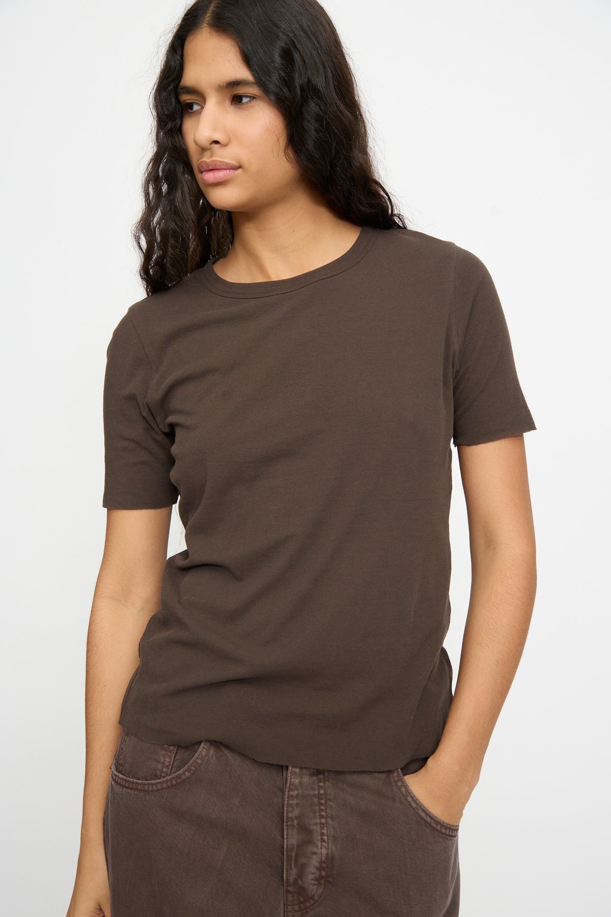 Person with long hair wearing the B Sides Double Tee in Walnut and Black and brown pants, standing against a plain white background.