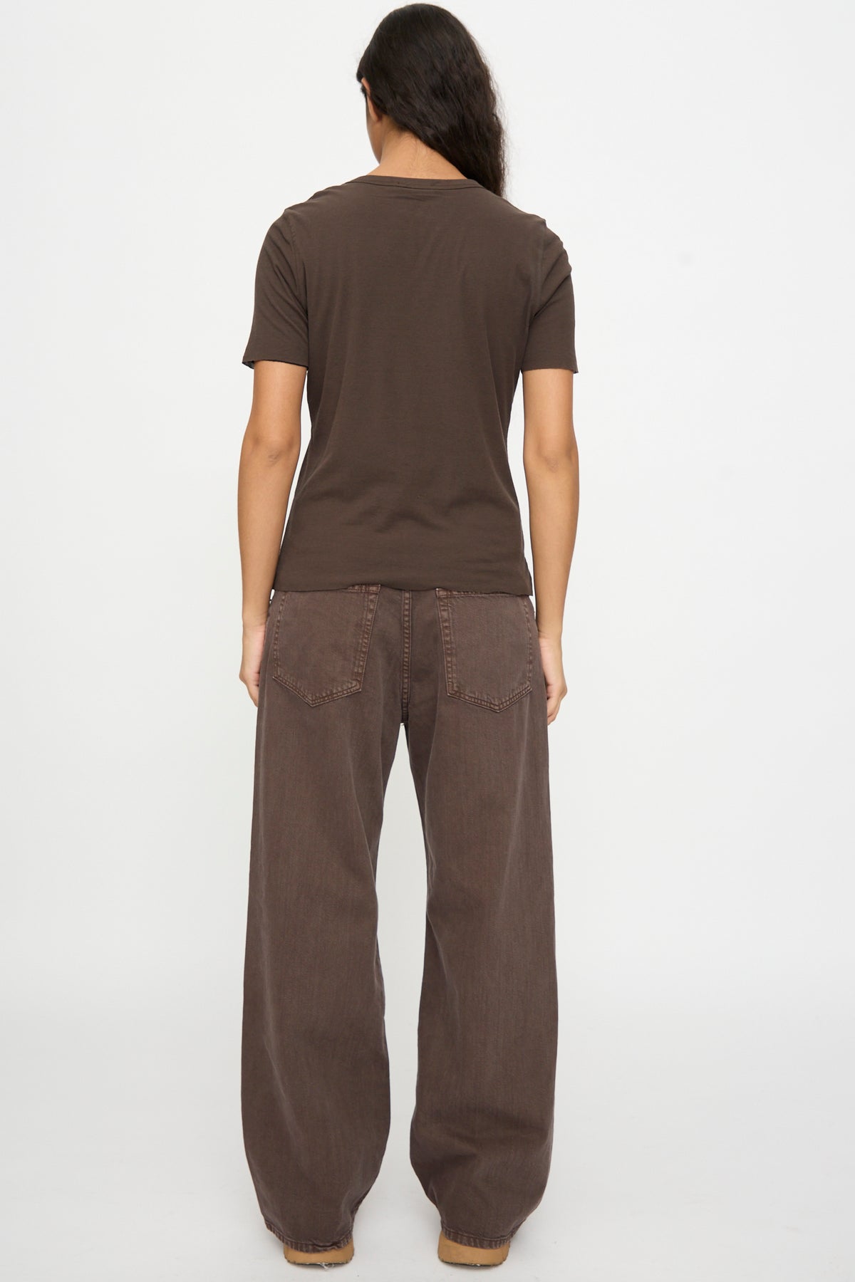 A person stands facing away in the B Sides Double Tee in Walnut and Black, paired with loose brown jeans against a plain white backdrop.