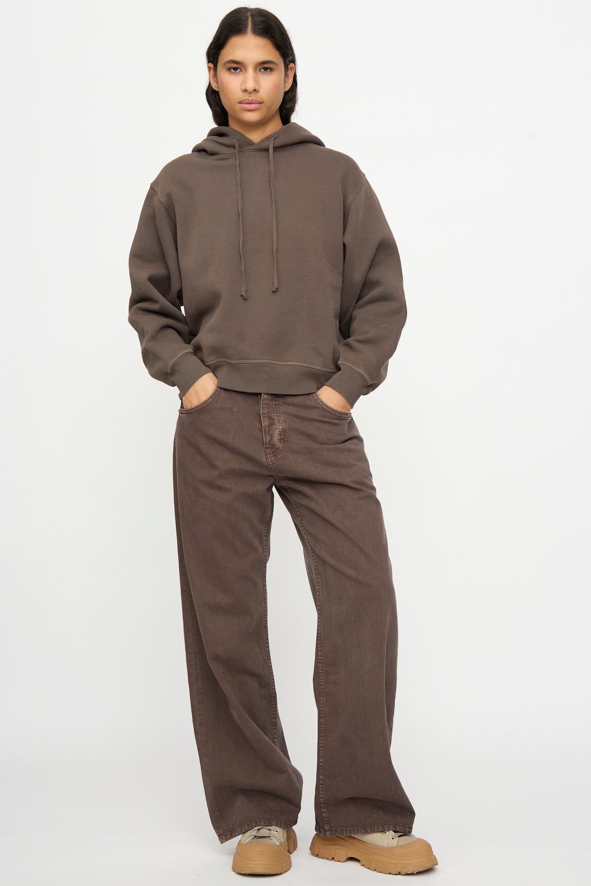 Person standing against a white background, wearing the B Sides Hoodie in Vintage Saddle Overdye, wide-leg pants, and tan shoes, with hands in pockets.