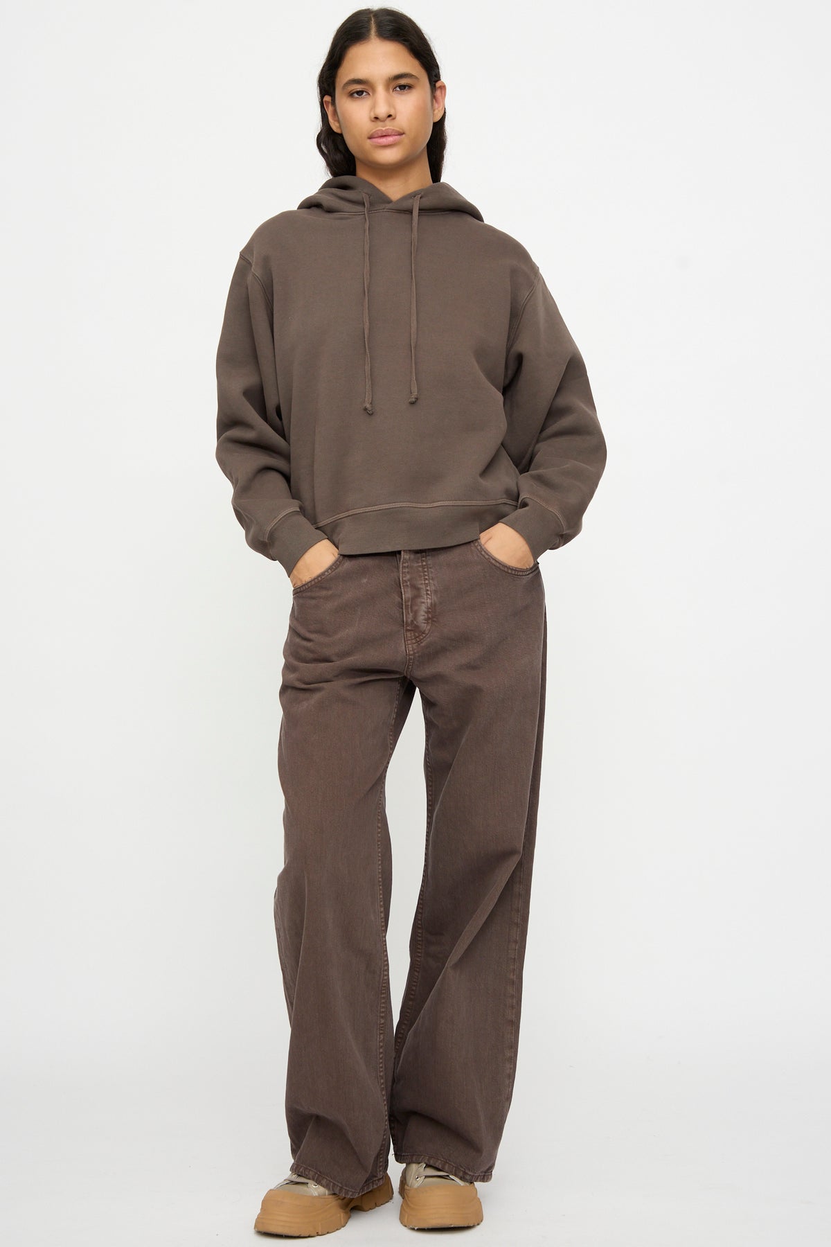 Model wears the B Sides Hoodie in Vintage Saddle Overdye, loose brown pants, and beige shoes, standing against a plain white background with hands in pockets.