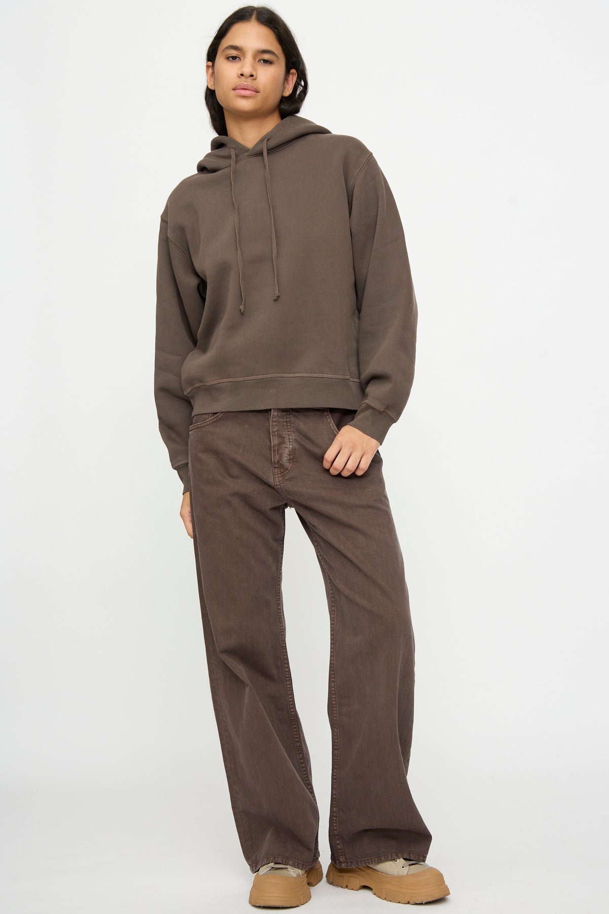 Person stands against a plain background wearing the B Sides Hoodie in Vintage Saddle Overdye, loose brown pants, tan shoes, and has one hand in a pocket.