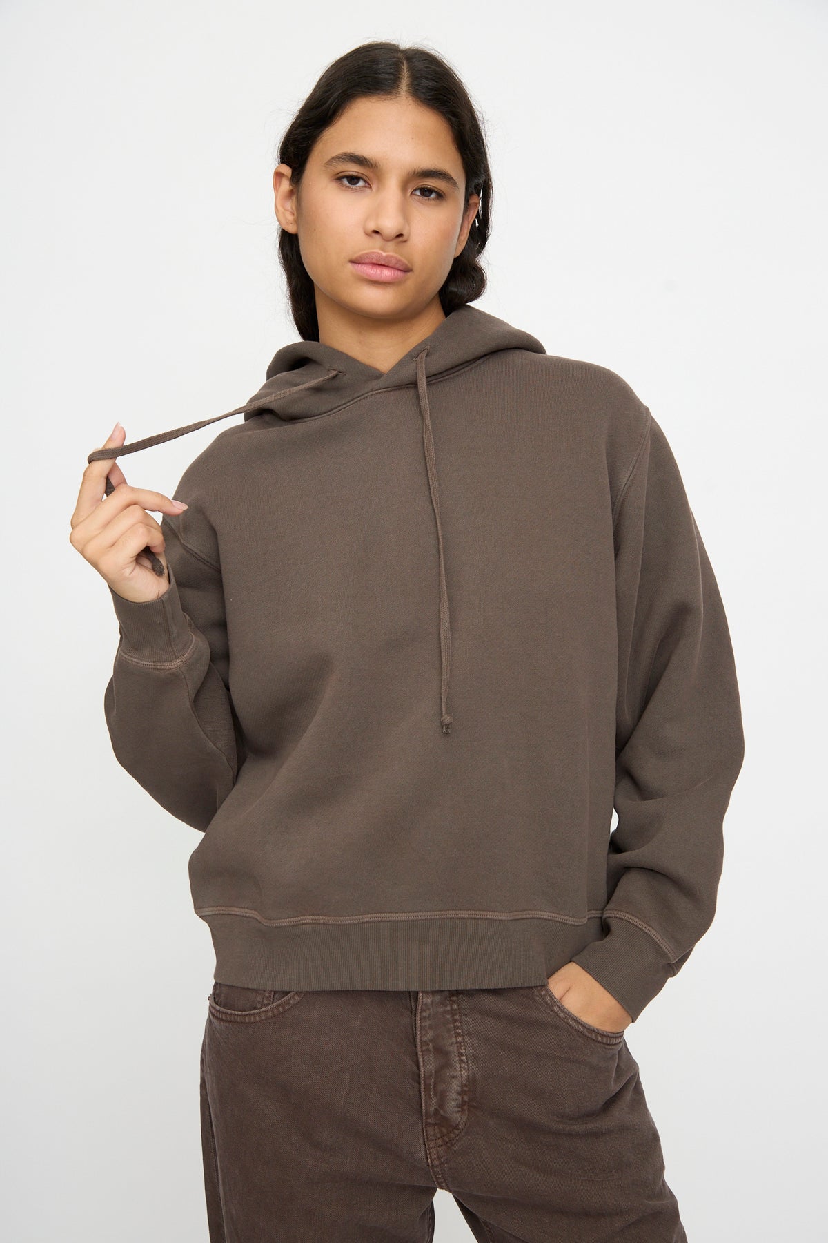 Model wears the B Sides Hoodie in Vintage Saddle Overdye with brown pants, one hand in pocket and the other holding a drawstring against a plain background.