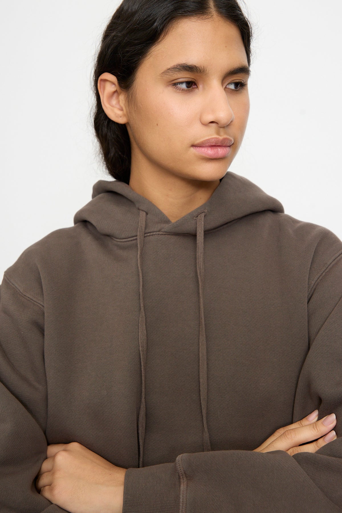 Person with long dark hair wears the B Sides Hoodie in Vintage Saddle Overdye, standing with arms crossed and looking to the side against a plain white background.