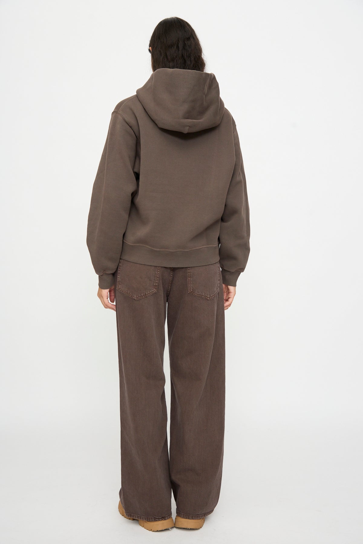 Person stands with their back to the camera wearing the B Sides Hoodie in Vintage Saddle Overdye, wide-leg brown jeans, and tan shoes against a plain white background.