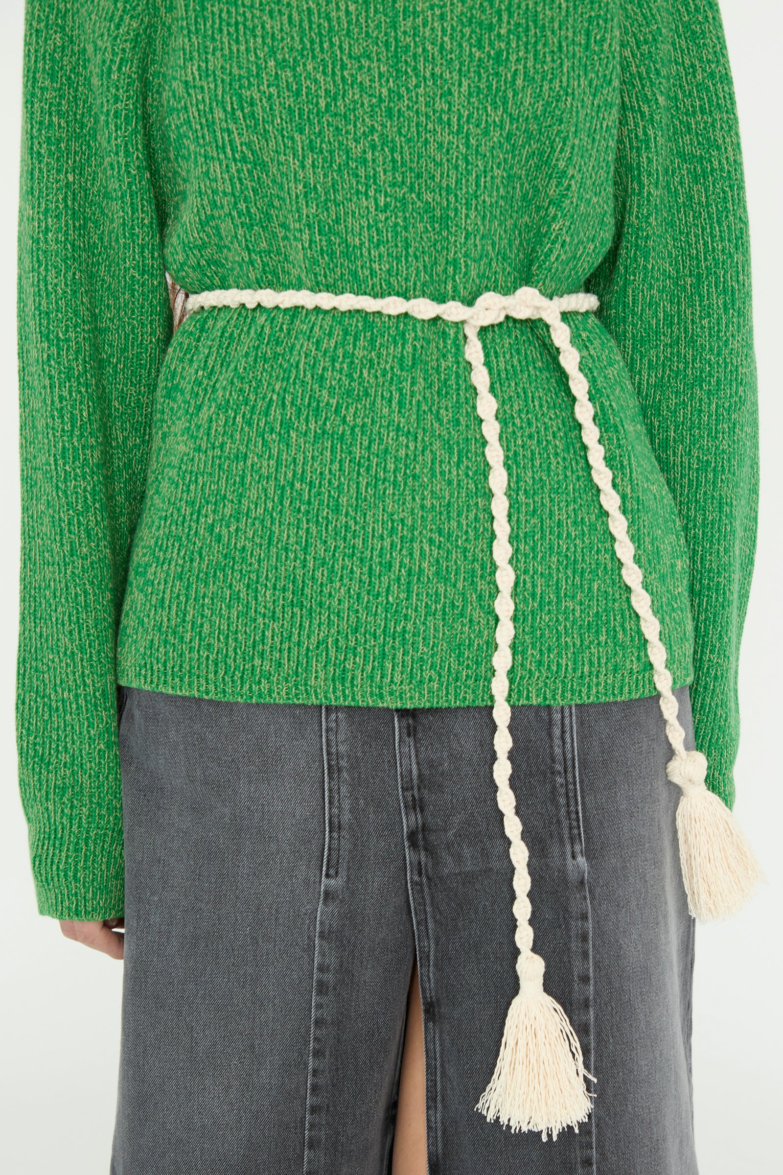 A person in a green sweater wears the B Sides Fran Macramé Belt in Ecru and pairs it with a high-waisted gray skirt featuring a back slit, made from Japanese cotton.