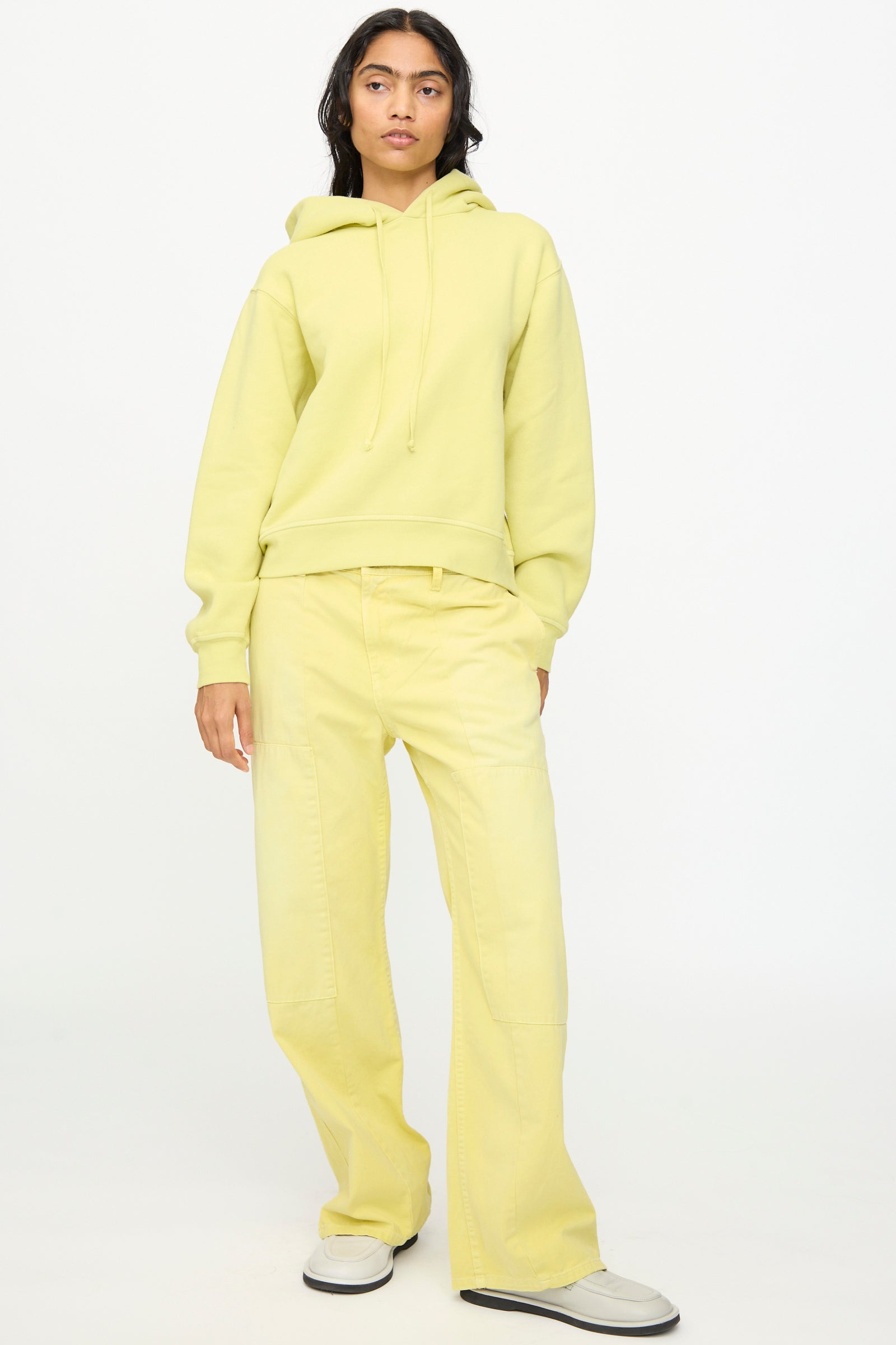 A person stands against a plain background wearing the B Sides Hoodie in Celery Overdye, styled with relaxed wide-leg pants and white slip-on shoes.