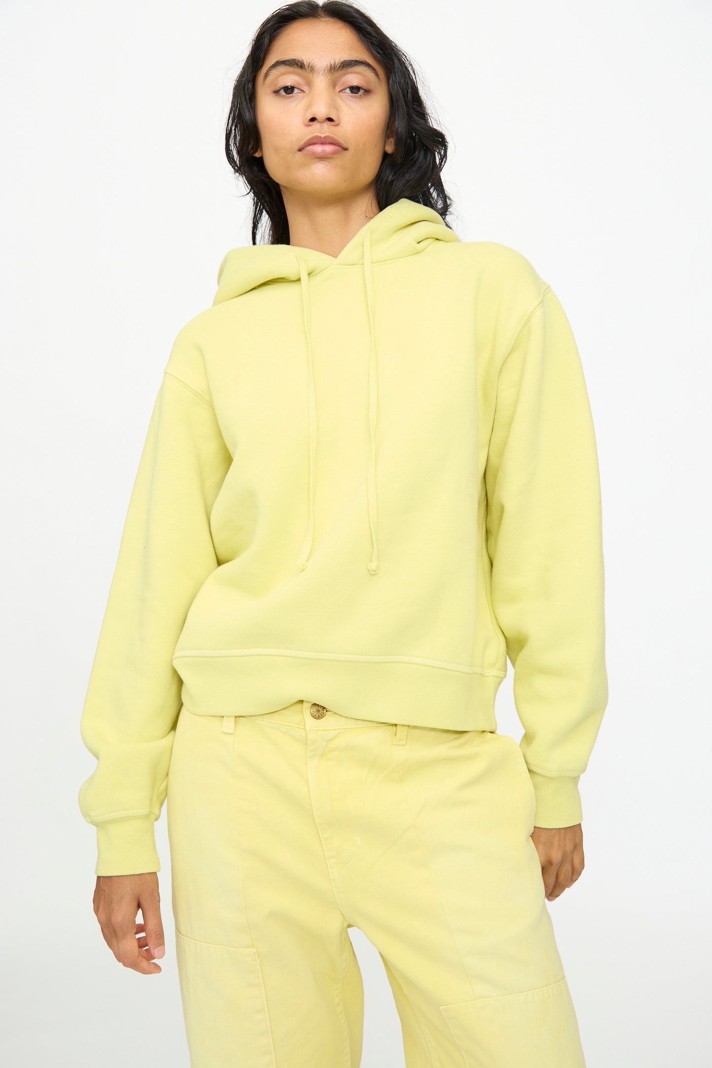 A person wearing the B Sides Hoodie in Celery Overdye—featuring a relaxed fit—and matching pale yellow pants, standing against a plain light background.
