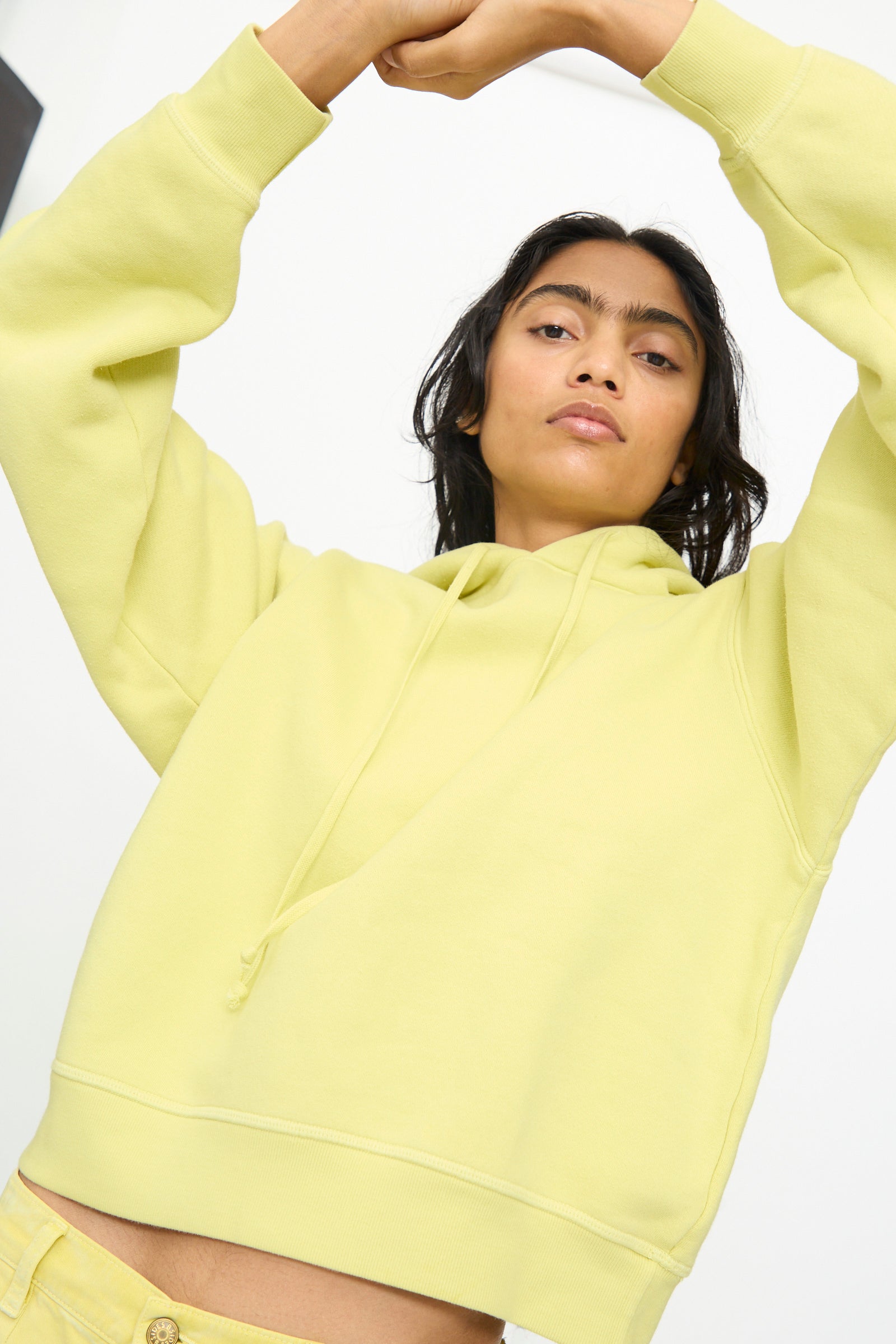 Person wearing the B Sides Hoodie in Celery Overdye, paired with yellow pants, posing with arms raised above head against a plain background.