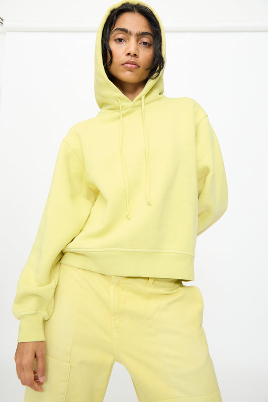 A person wearing the B Sides Hoodie in Celery Overdye—a light yellow, relaxed fit hoodie made from overdyed cotton and crafted in the USA—paired with matching pants, stands against a plain white background.