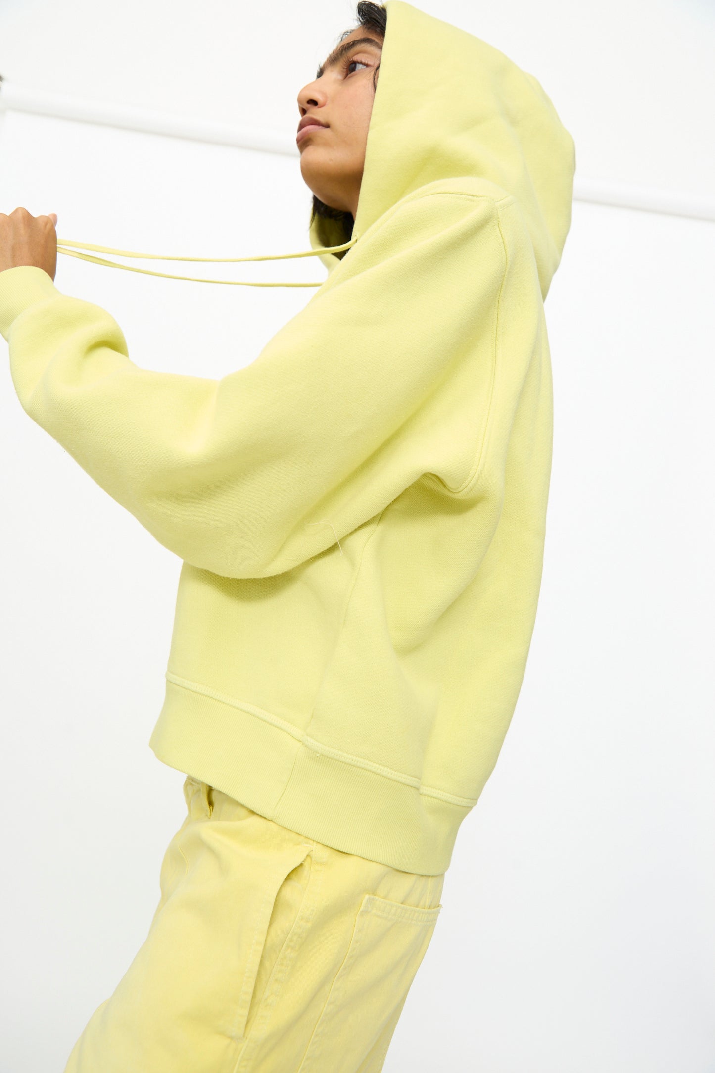 A person wearing the B Sides Hoodie in Celery Overdye and matching pants stands against a white background, holding the hoodie strings and looking upward.