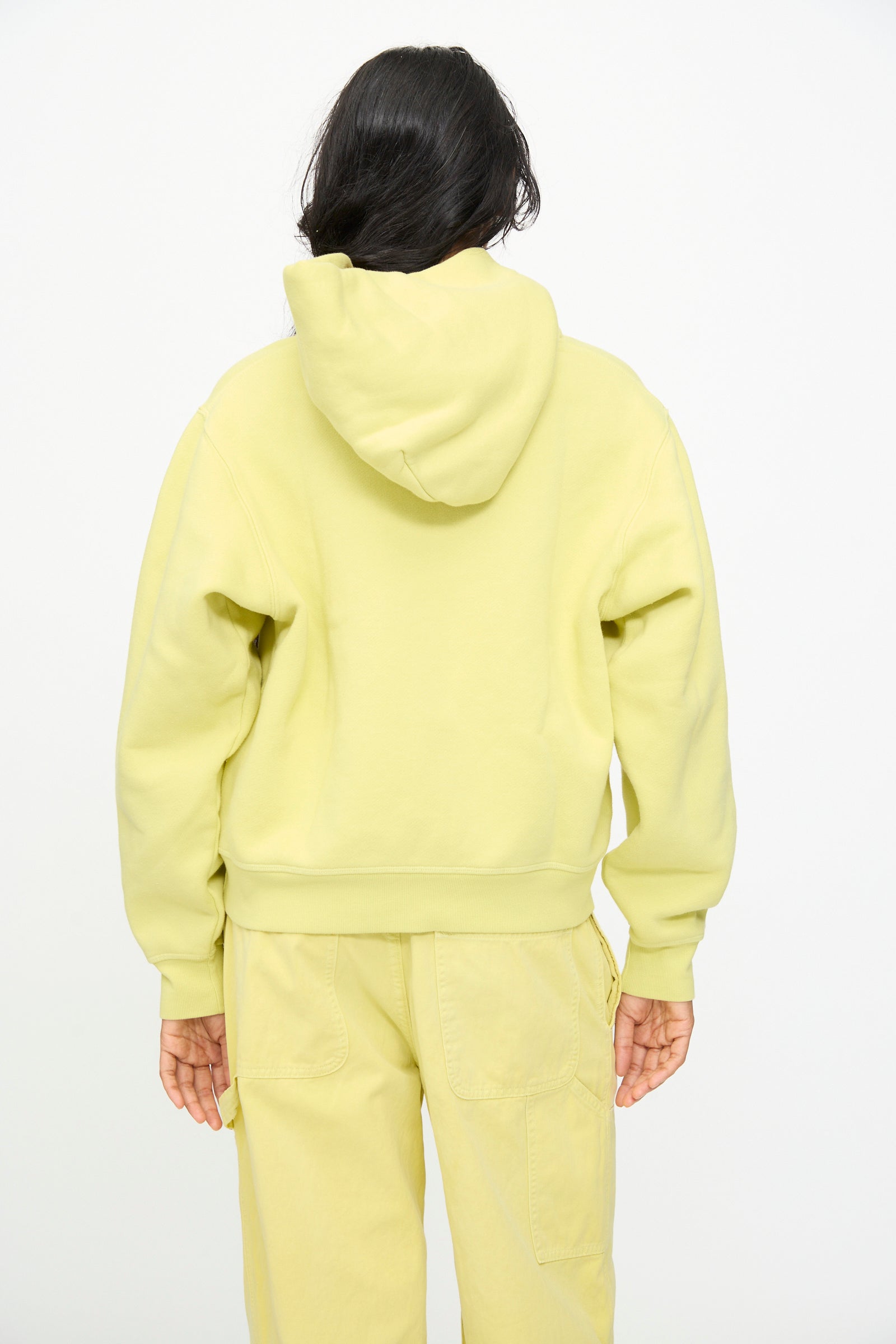 A person stands with their back to the camera, wearing the B Sides Hoodie in Celery Overdye and matching pants against a plain white background.