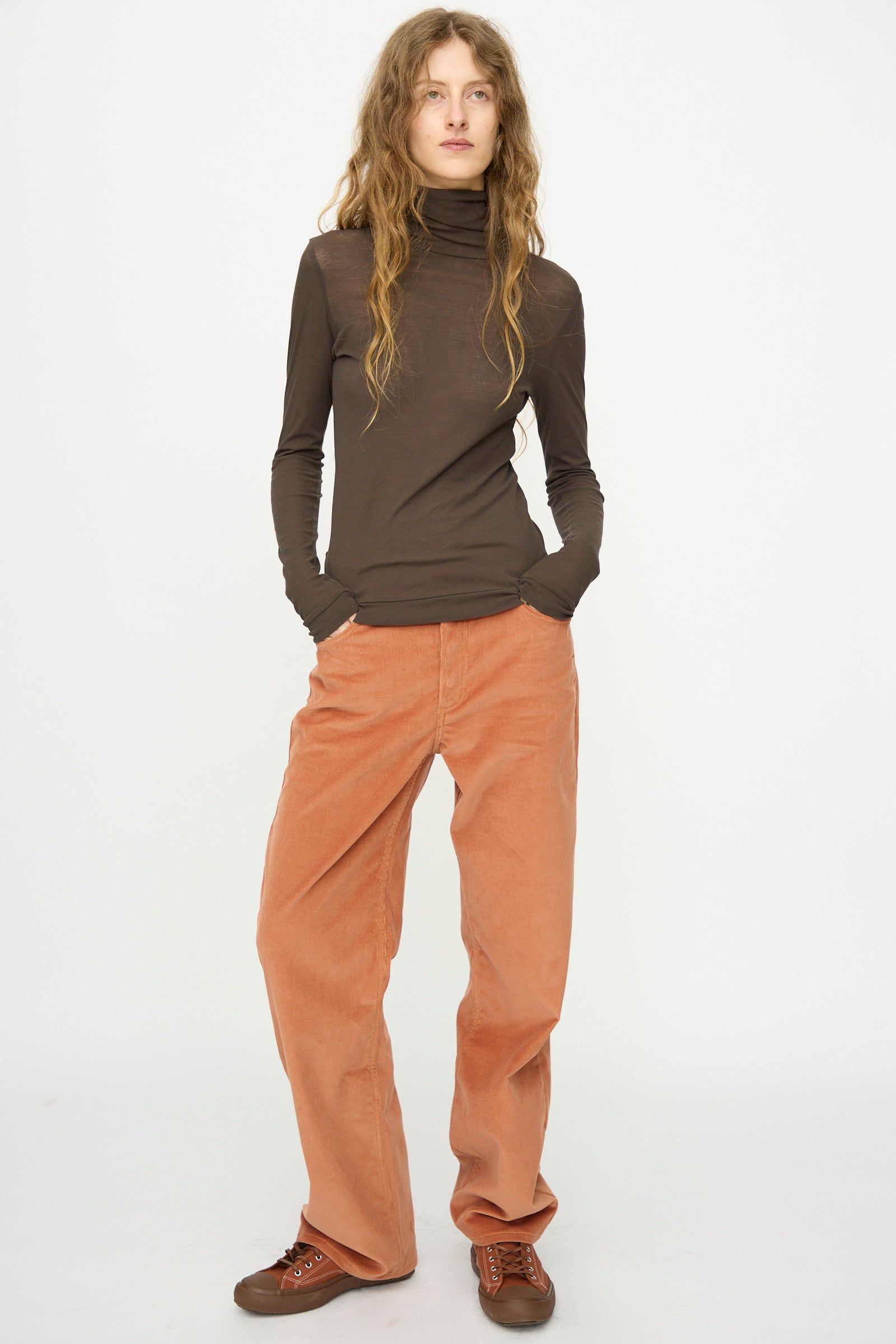 A person stands against a plain white background wearing the B Sides Jana Turtleneck in Olive Drab, loose peach corduroy pants, and brown sneakers, hands in pockets.