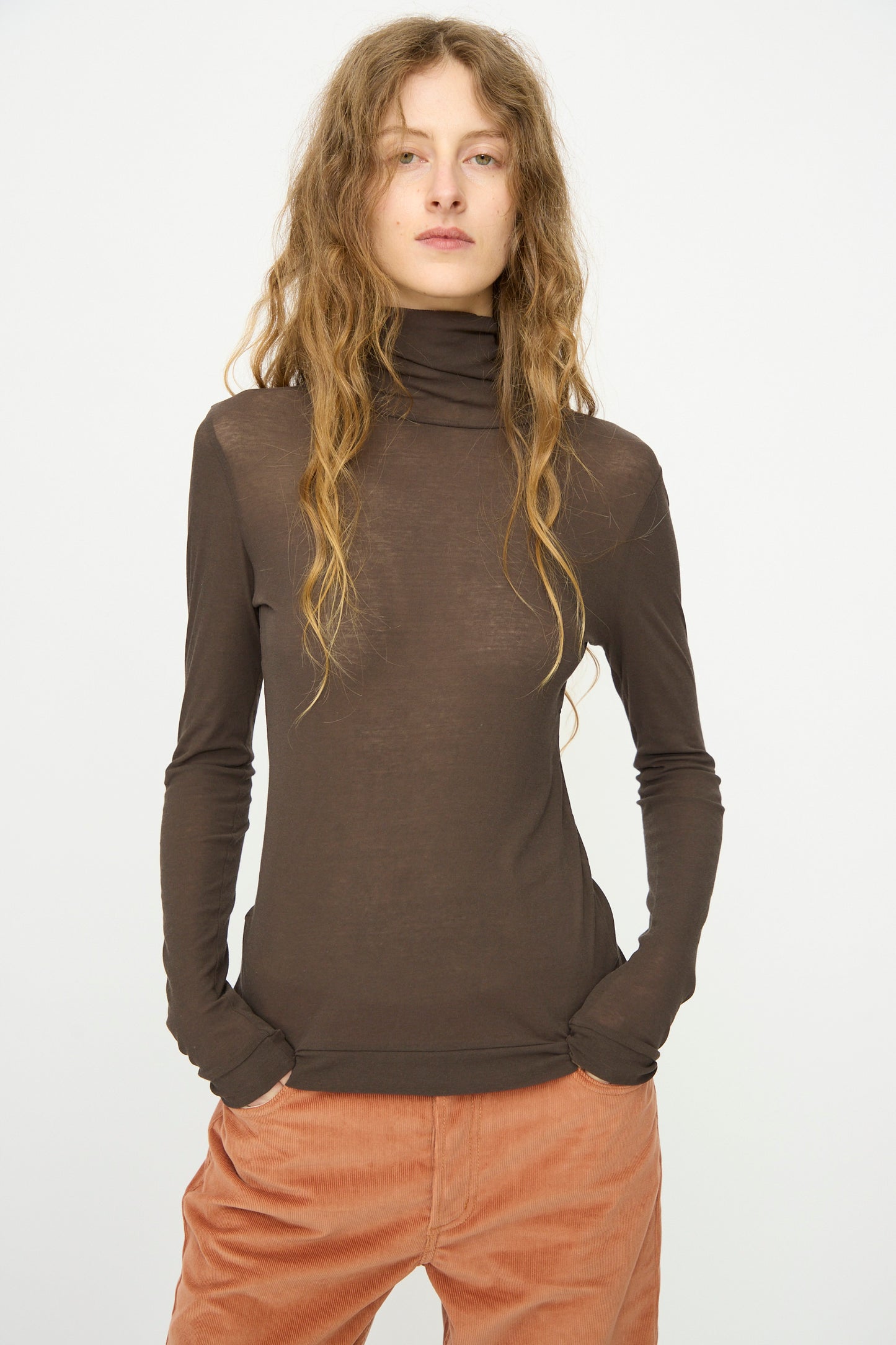 A person with long wavy hair wears the B Sides Jana Turtleneck in Olive Drab and rust-colored pants, standing with hands in pockets against a plain white background.