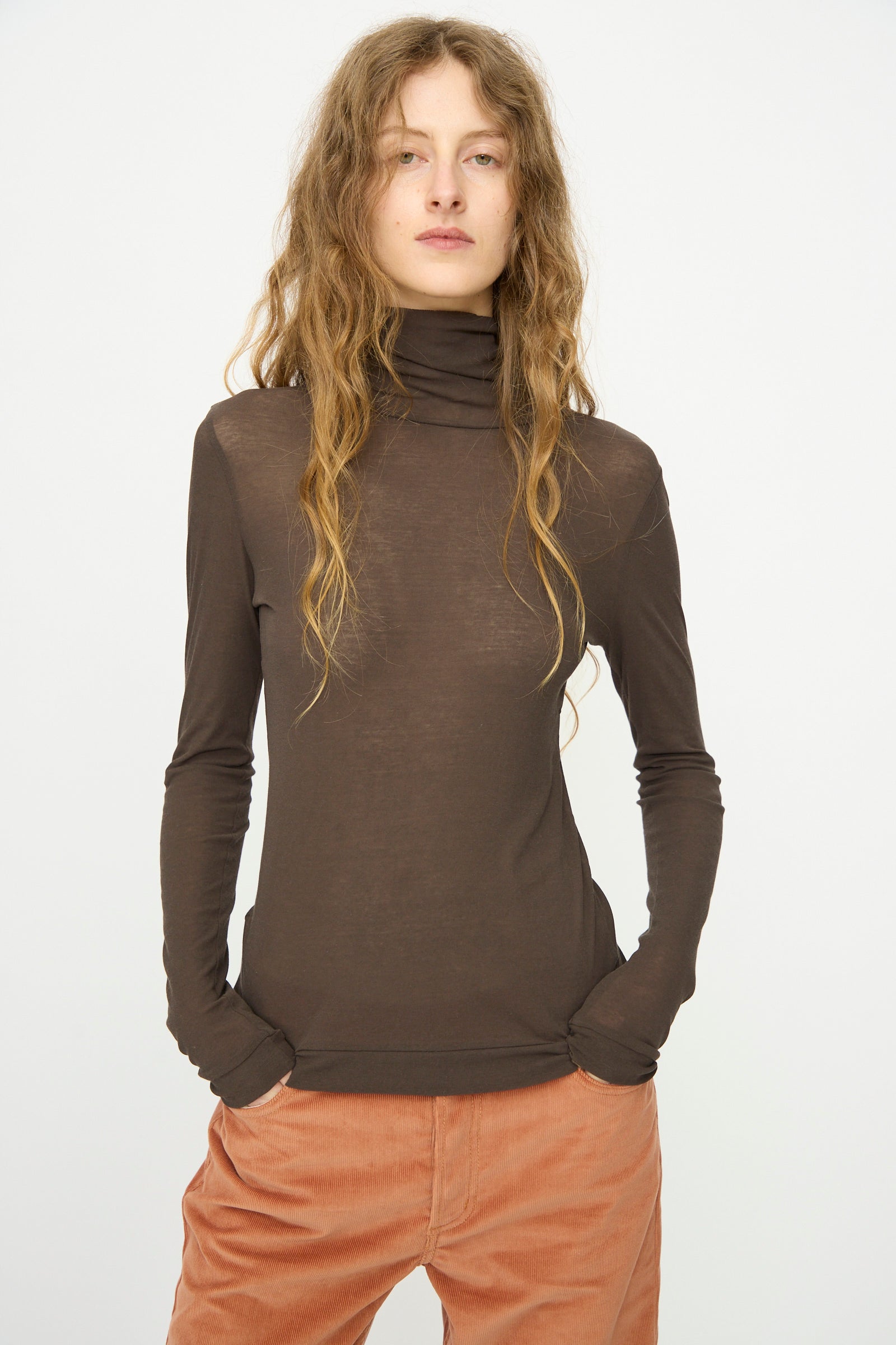 A person with long wavy hair wears the B Sides Jana Turtleneck in Olive Drab and rust-colored pants, standing with hands in pockets against a plain white background.