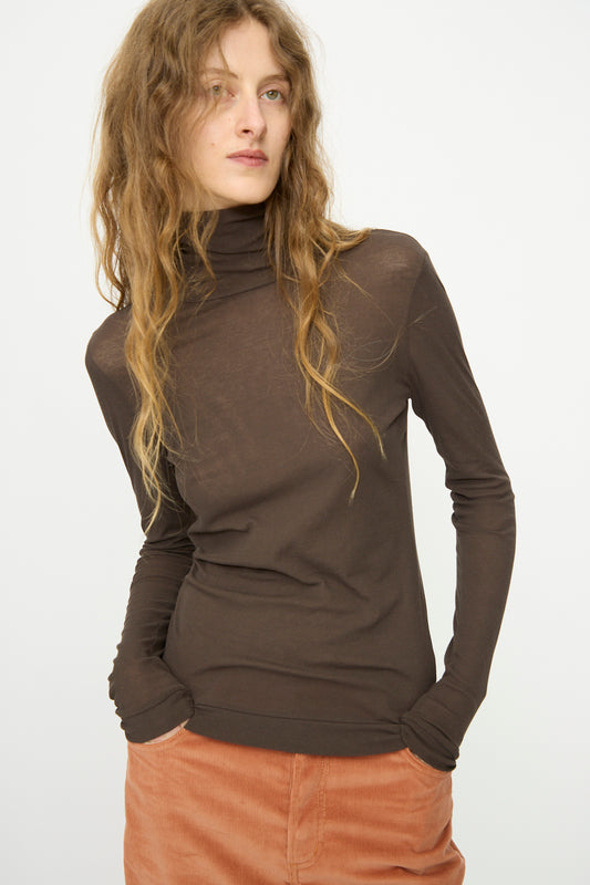 Person with long wavy hair wears the B Sides Jana Turtleneck in Olive Drab (slim fit, superfine cotton rib) and peach pants, standing hands-in-pockets against a plain light background.