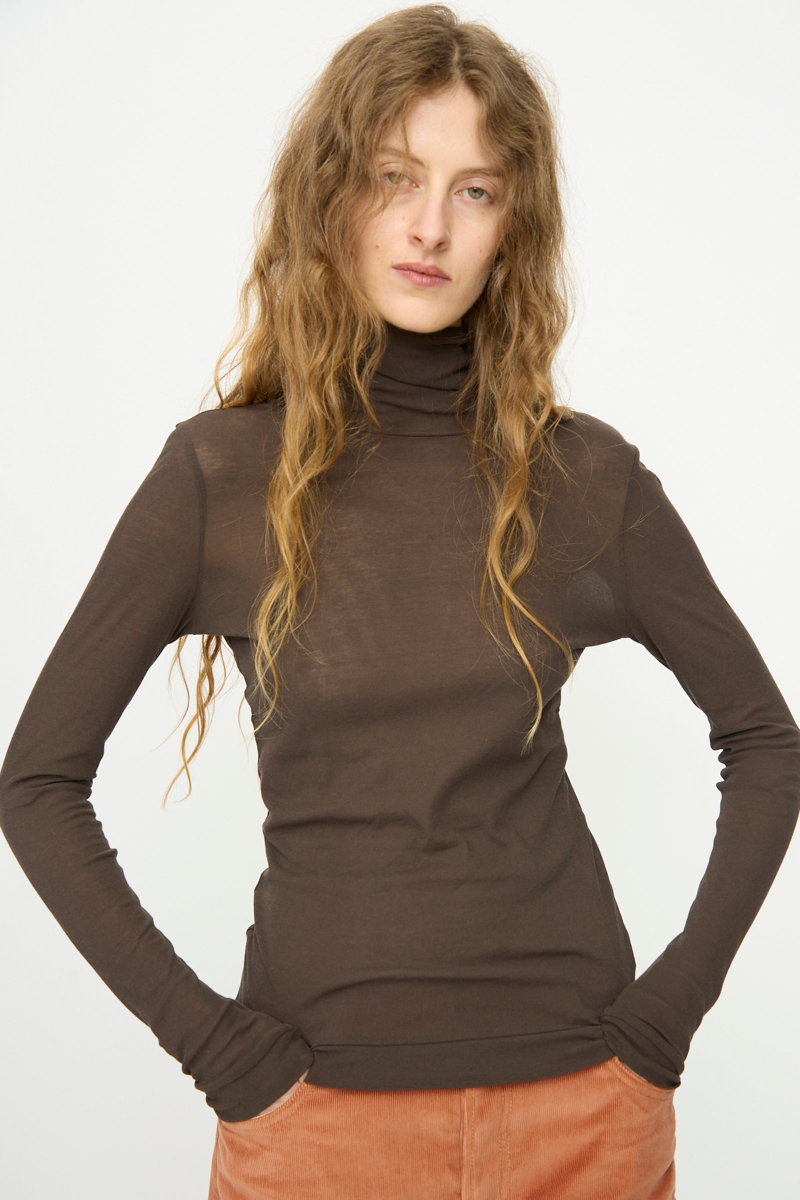 A person with long wavy hair wears the B Sides Jana Turtleneck in Olive Drab, paired with light brown corduroy pants, standing against a plain white background.
