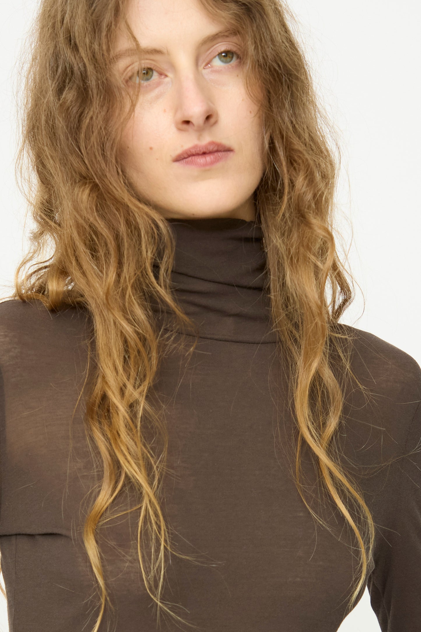 A person with long wavy hair wears the B Sides Jana Turtleneck in Olive Drab, a slim fit, sheer top crafted from superfine cotton rib, and looks slightly upward against a plain background.