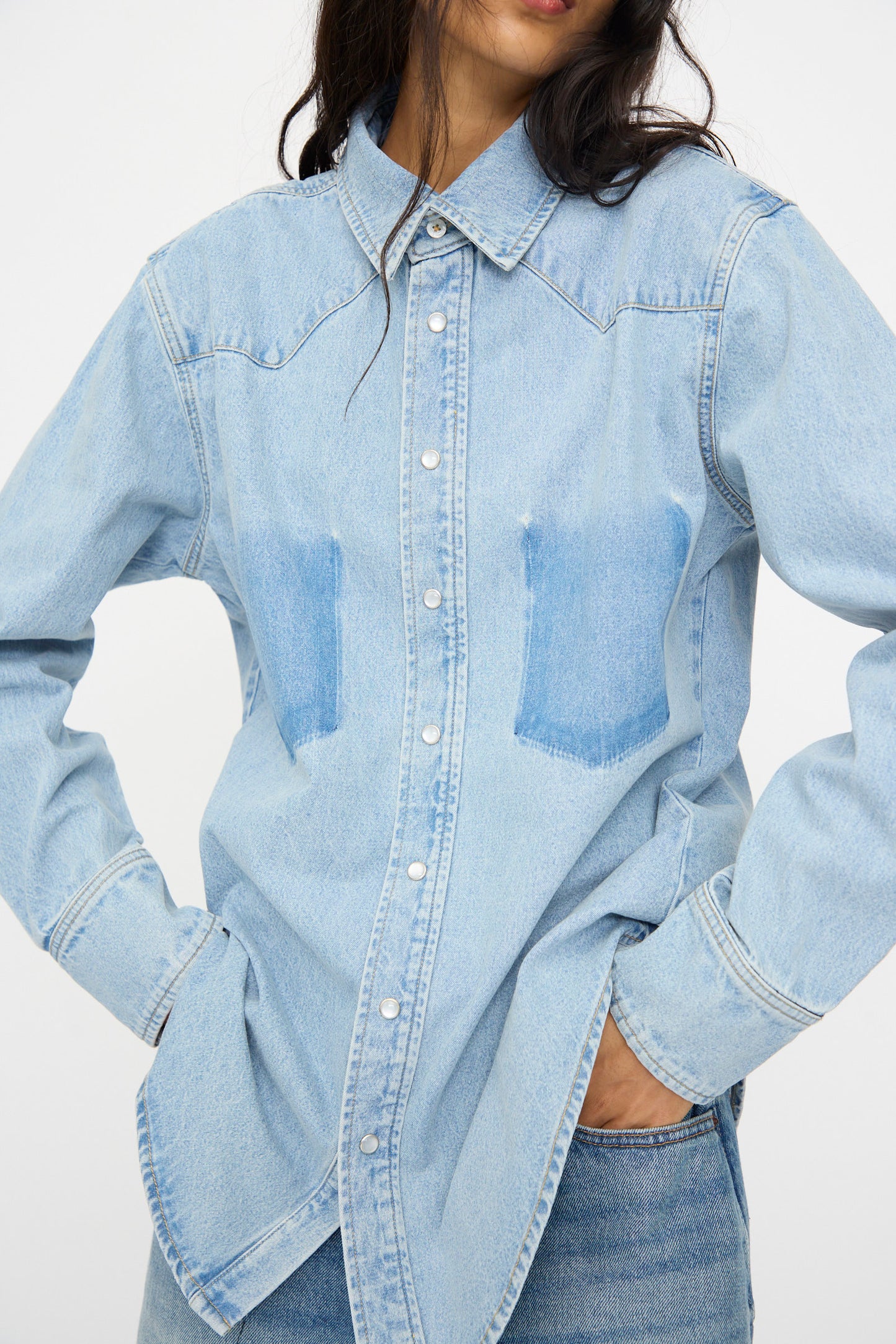 A person wears the B Sides Liv Western Shirt in Park Wash, a relaxed fit, light blue denim top, with hands in jean pockets while standing against a plain white background.