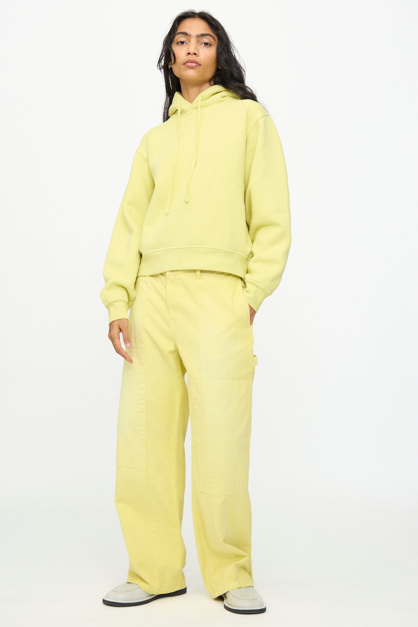 Wearing B Sides’ Painter Pant in Faded Celery Overdye, styled with a light yellow hoodie and white sneakers, the person stands against a plain white background.