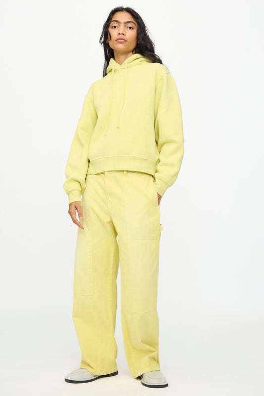 Wearing B Sides’ Painter Pant in Faded Celery Overdye, styled with a light yellow hoodie and white sneakers, the person stands against a plain white background.