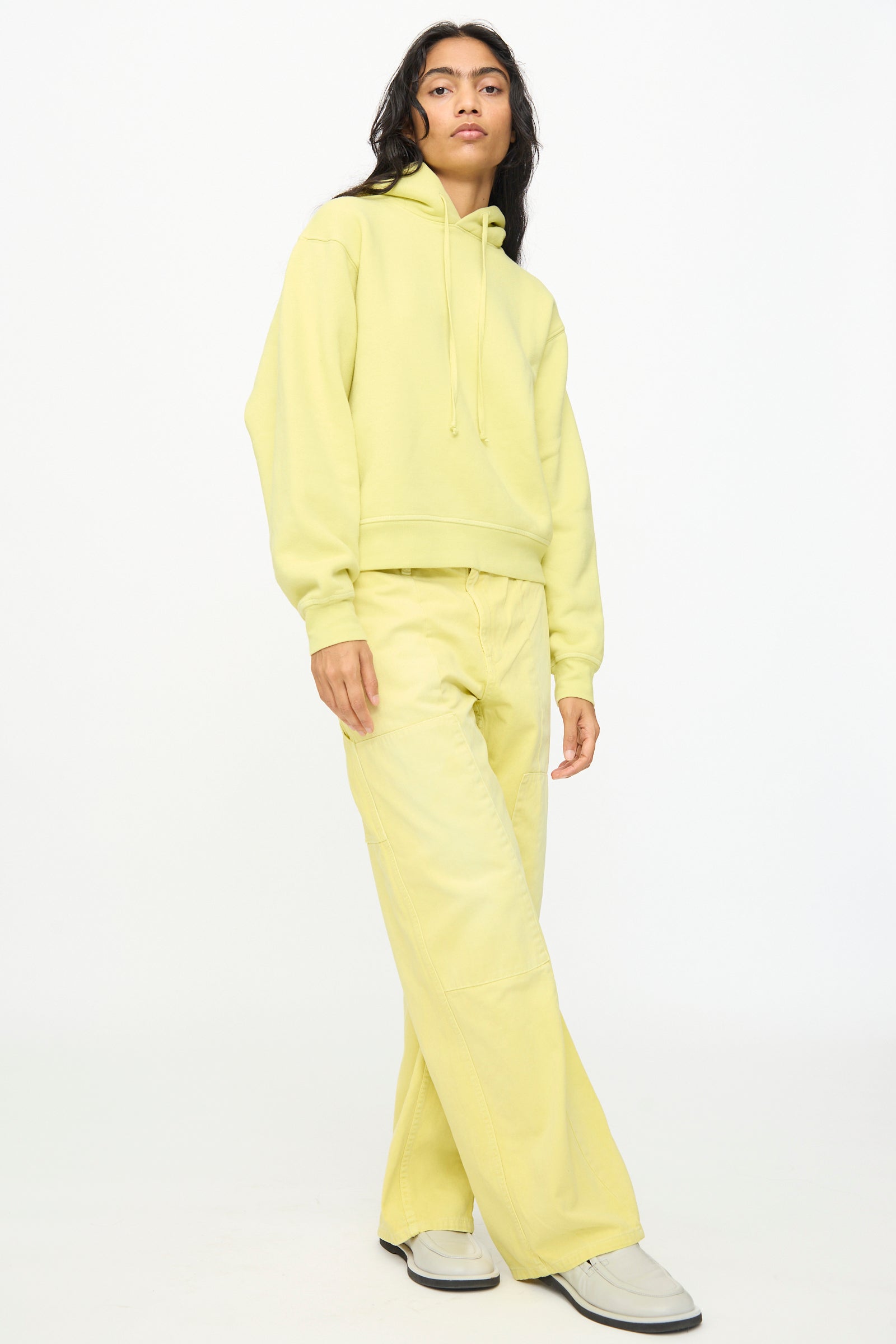 Person wearing a light yellow relaxed fit hoodie and B Sides' Painter Pant in Faded Celery Overdye, paired with white shoes, standing against a plain white background.