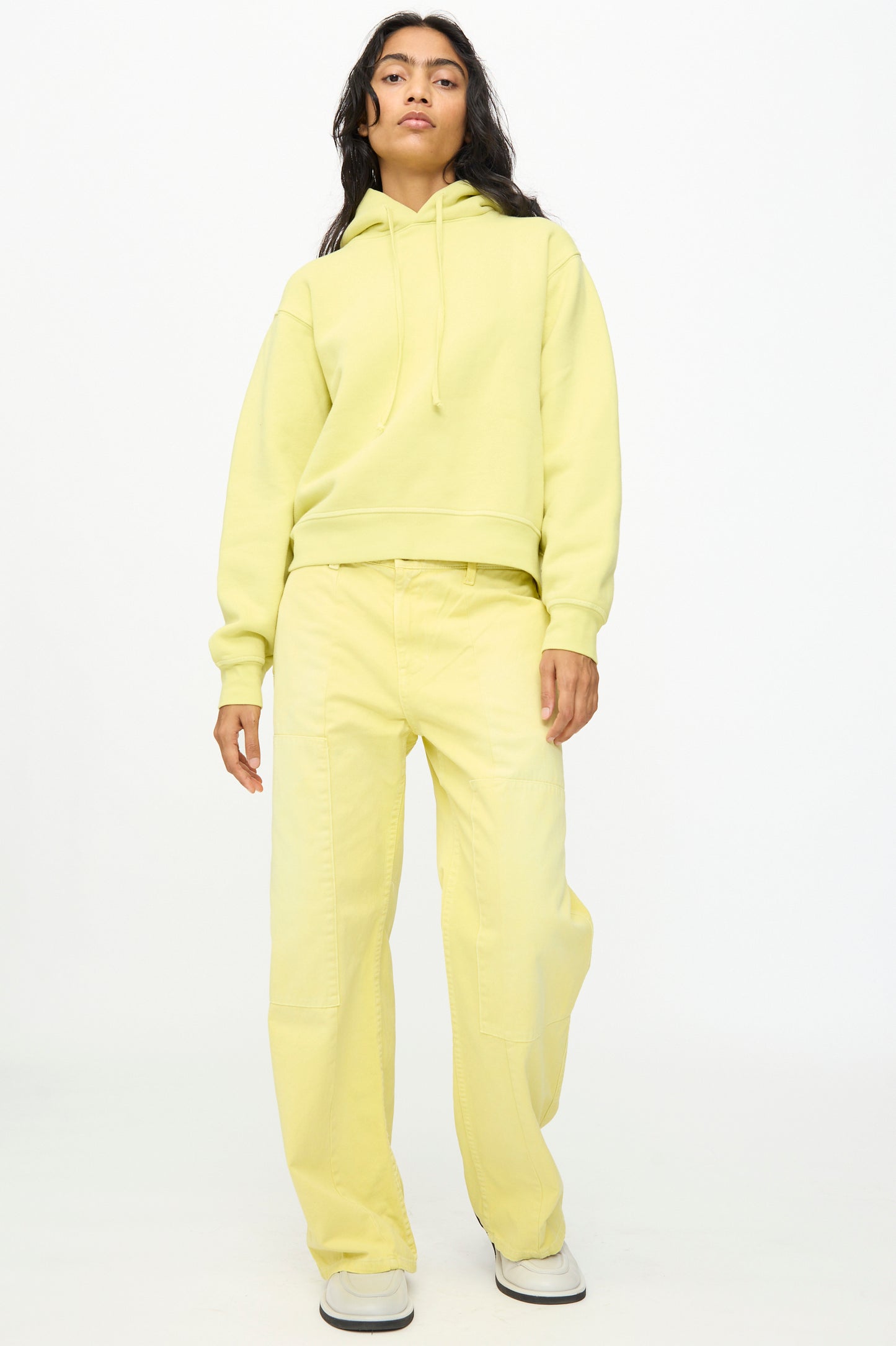 A person stands against a plain background wearing a yellow hoodie, B Sides Painter Pant in Faded Celery Overdye, and white shoes.