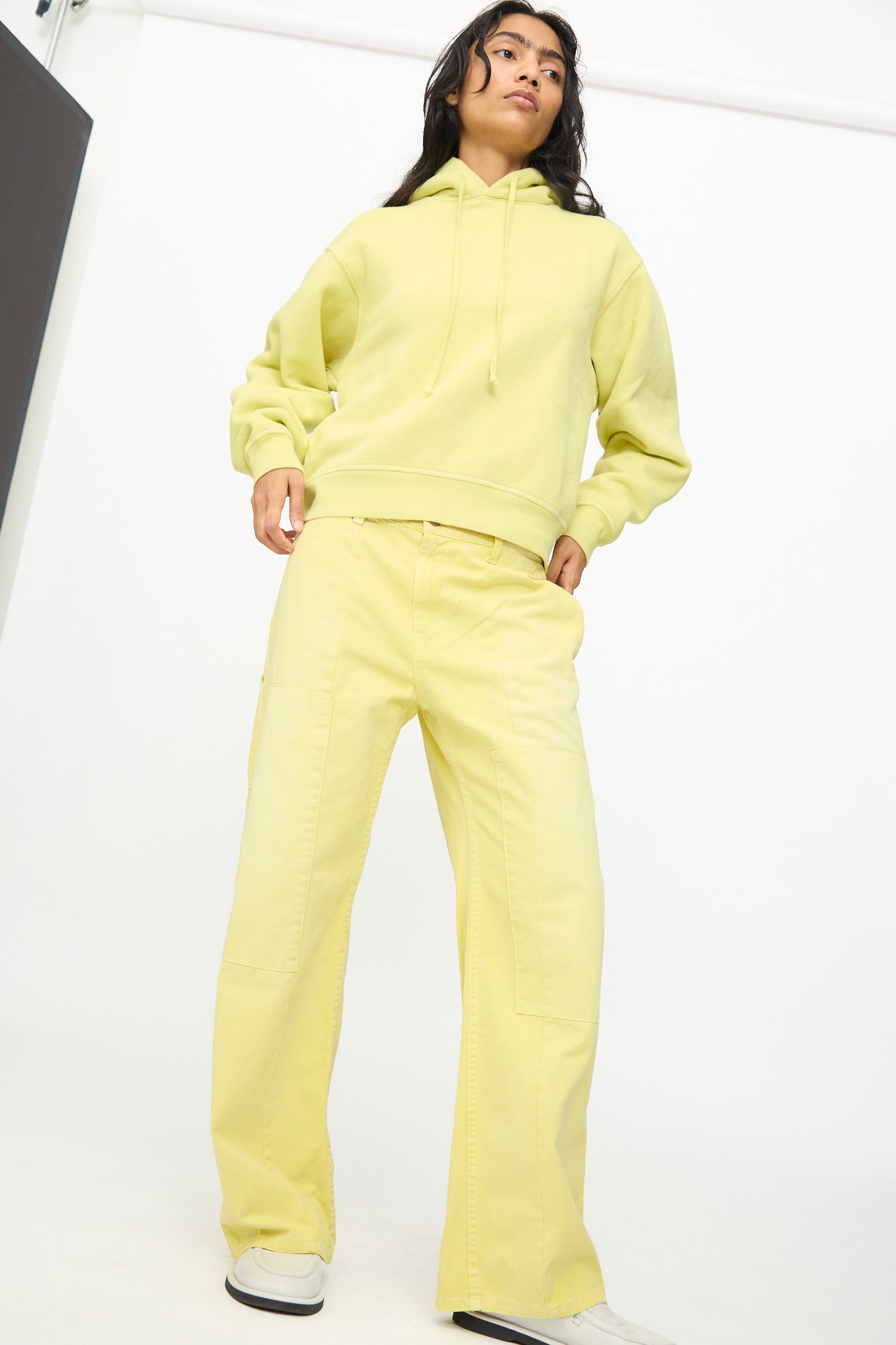 A person wearing the B Sides Painter Pant in Faded Celery Overdye, styled with a light yellow hoodie, stands against a plain white background looking slightly upward.
