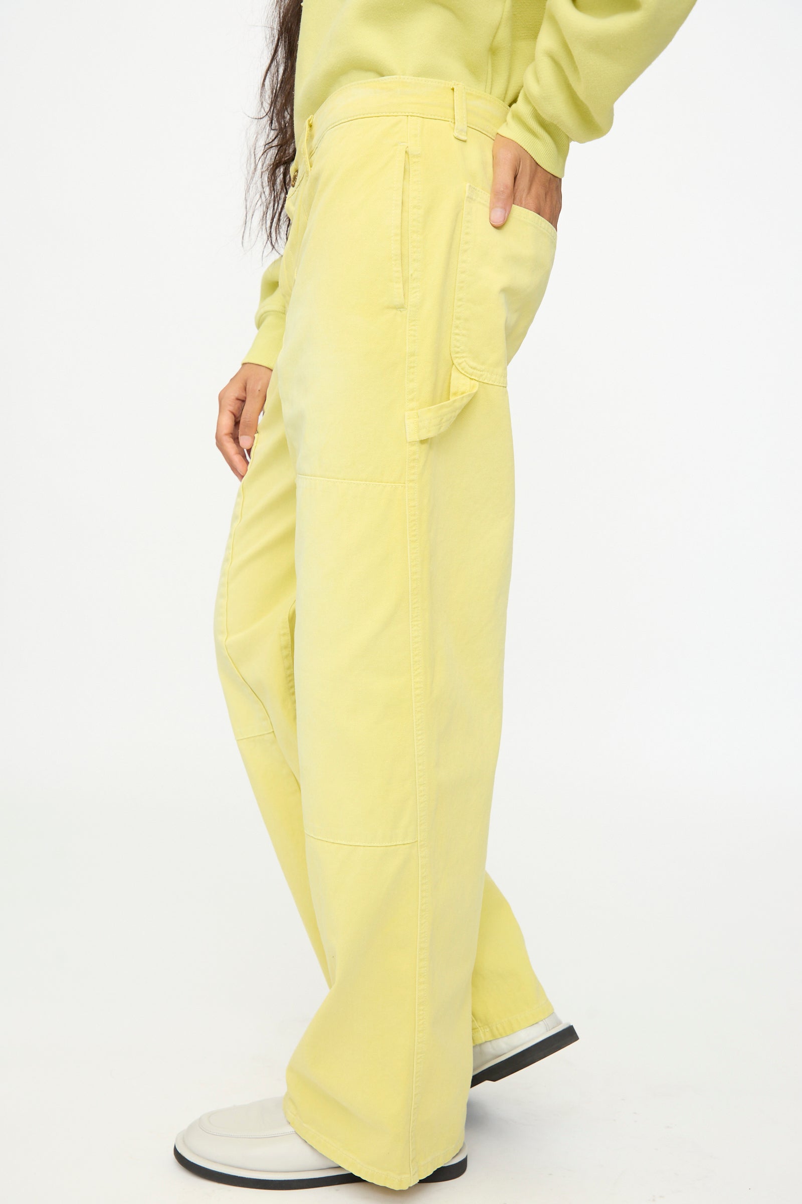 A person stands sideways against a plain background, wearing the B Sides Painter Pant in Faded Celery Overdye—a relaxed fit yellow cargo pant—paired with a yellow sweatshirt and white shoes, with one hand in their pocket.