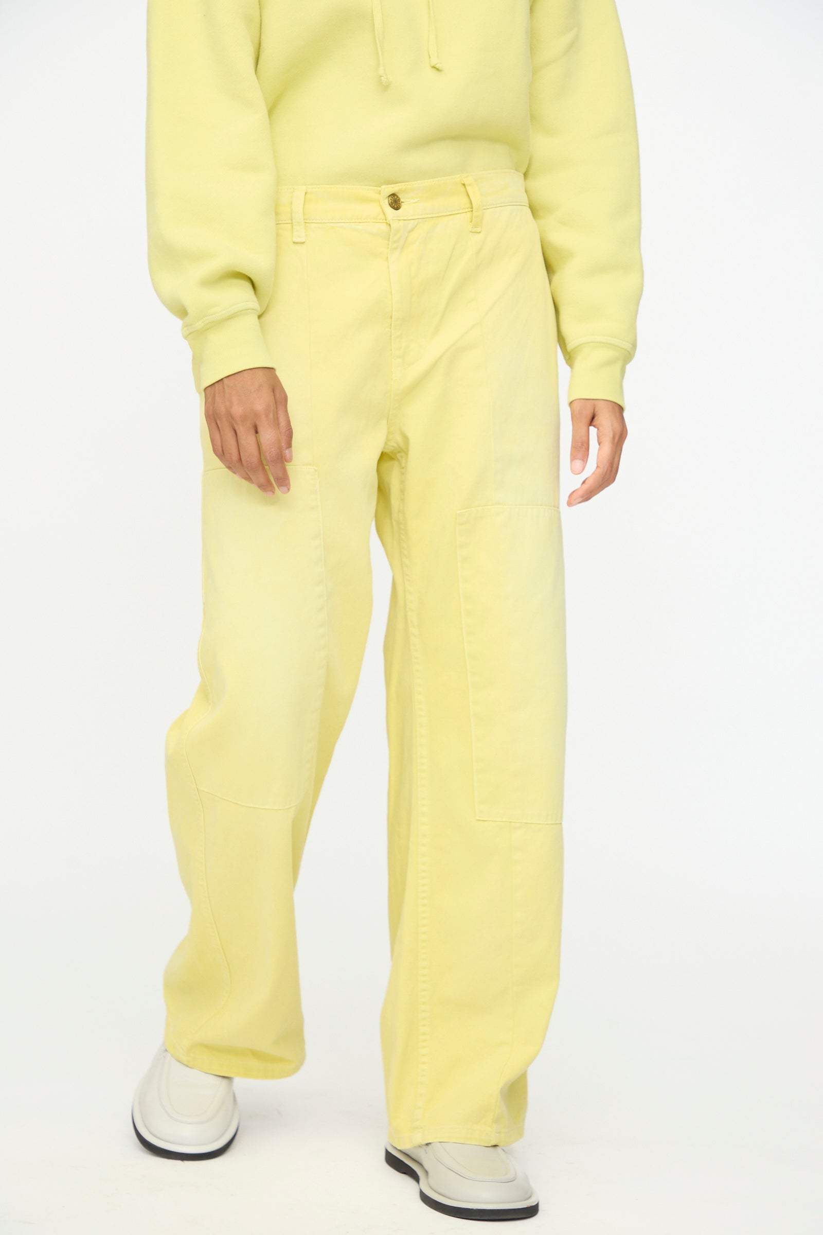 A person wearing a pale yellow hoodie and B Sides Painter Pant in Faded Celery Overdye with white shoes, standing against a plain white background.