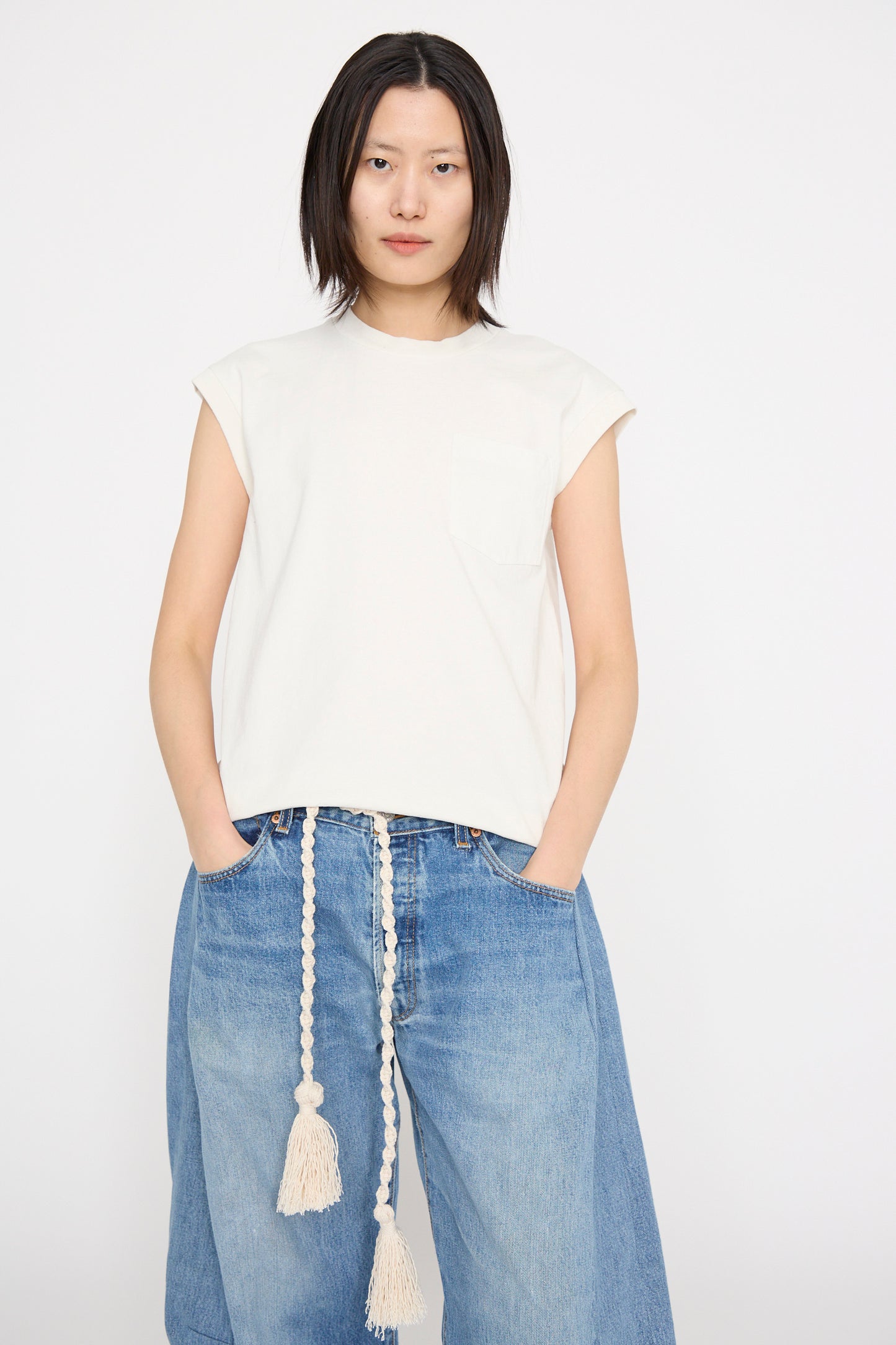 Wearing a Pocket Tank in Snow White by B Sides and blue jeans with a long braided belt, a model stands against a plain background, facing forward. 