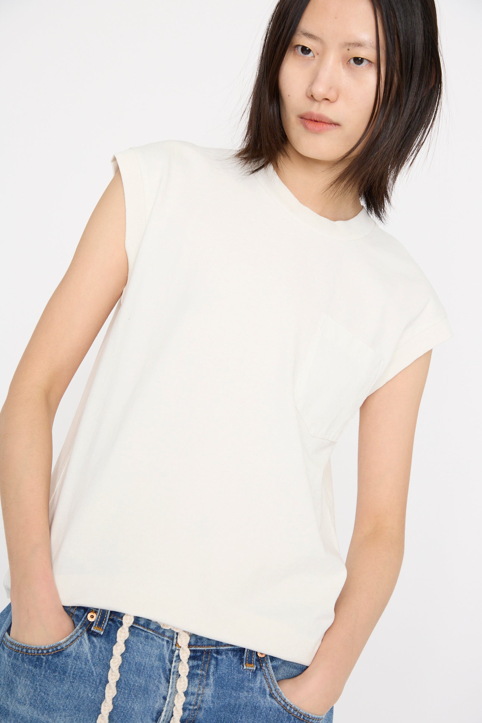 With shoulder-length hair, a person wears the Pocket Tank in Snow White by B Sides with blue jeans against a simple backdrop.