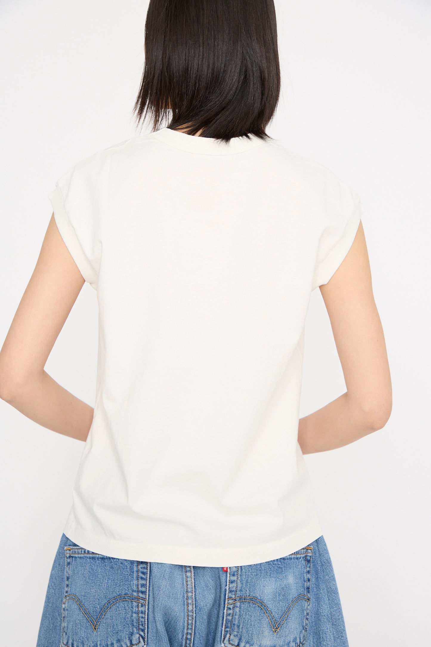 A rear view of a person with short black hair is shown, wearing a Pocket Tank in Snow White by B Sides and blue jeans against a plain white background.