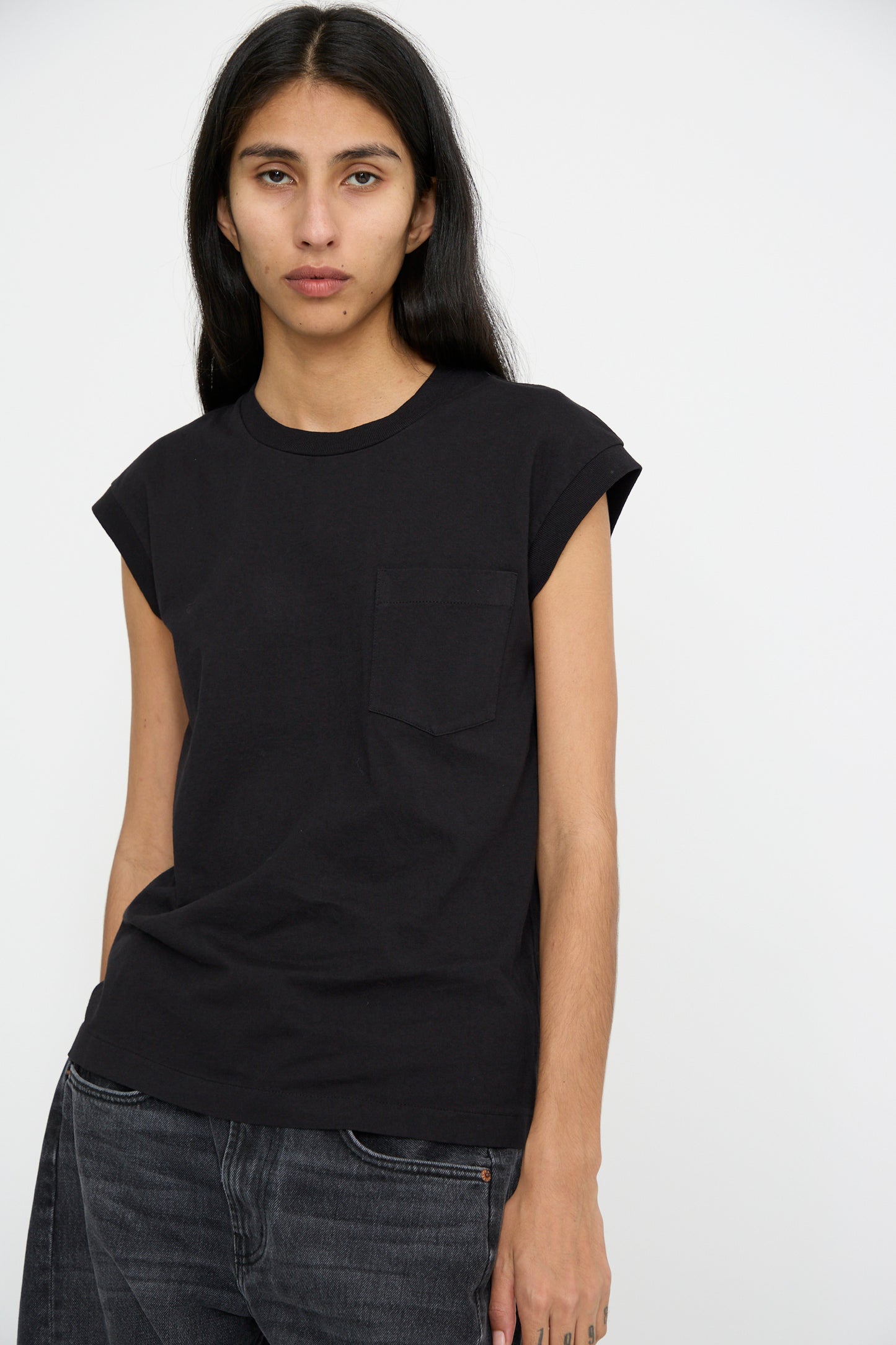 A person with long black hair poses against a plain background wearing the B Sides Pocket Tank in Stil Black, which features a single front pocket, and looks forward.