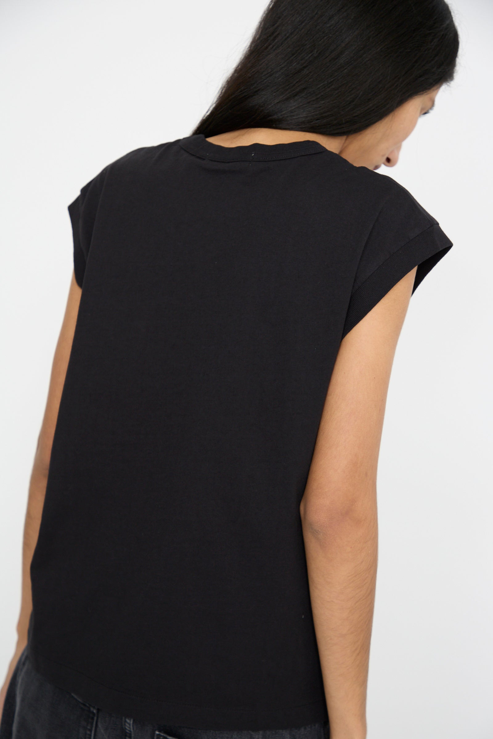 A person with long dark hair is facing away, wearing the Pocket Tank in Stil Black by B Sides, featuring a black sleeveless design with a loose knit jersey texture.