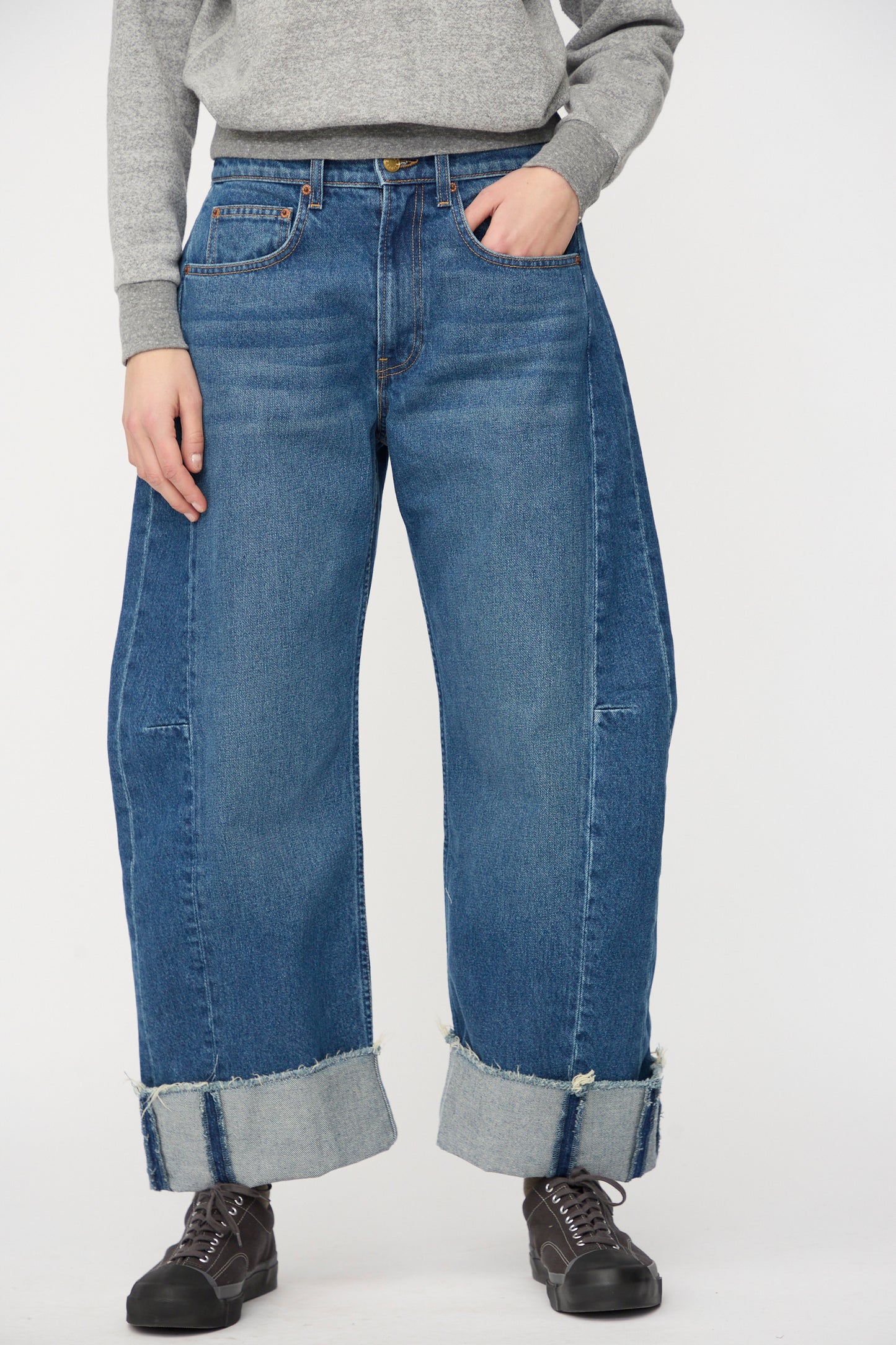 A person stands against a plain background wearing a grey sweatshirt, black lace-up shoes, and B Sides Relaxed Cuffed Lasso Jean in Cate Wash featuring wide legs with large cuffs.