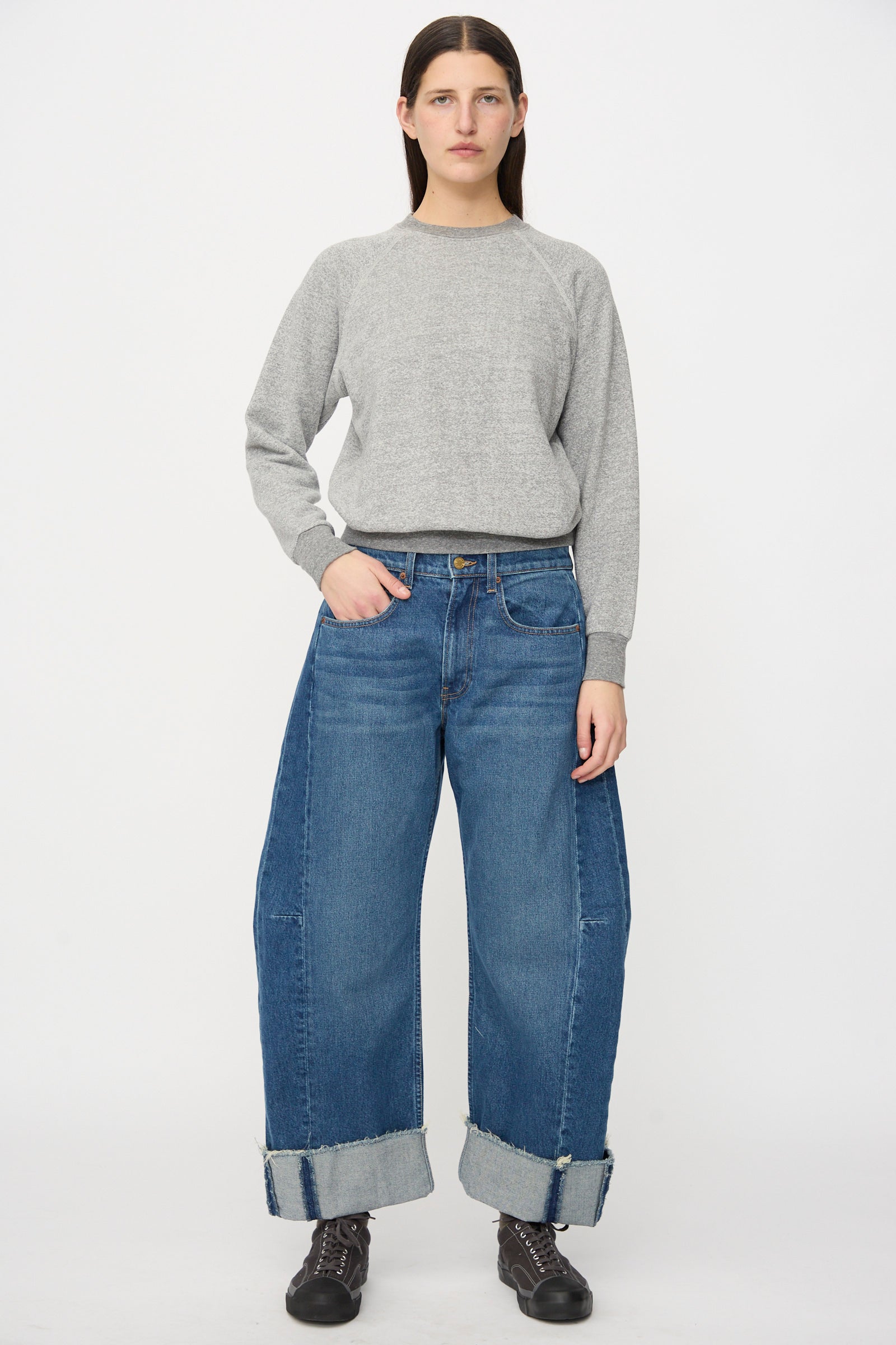 A person stands facing forward in a grey sweater, B Sides Relaxed Cuffed Lasso Jean in Cate Wash, and black boots against a plain white background.