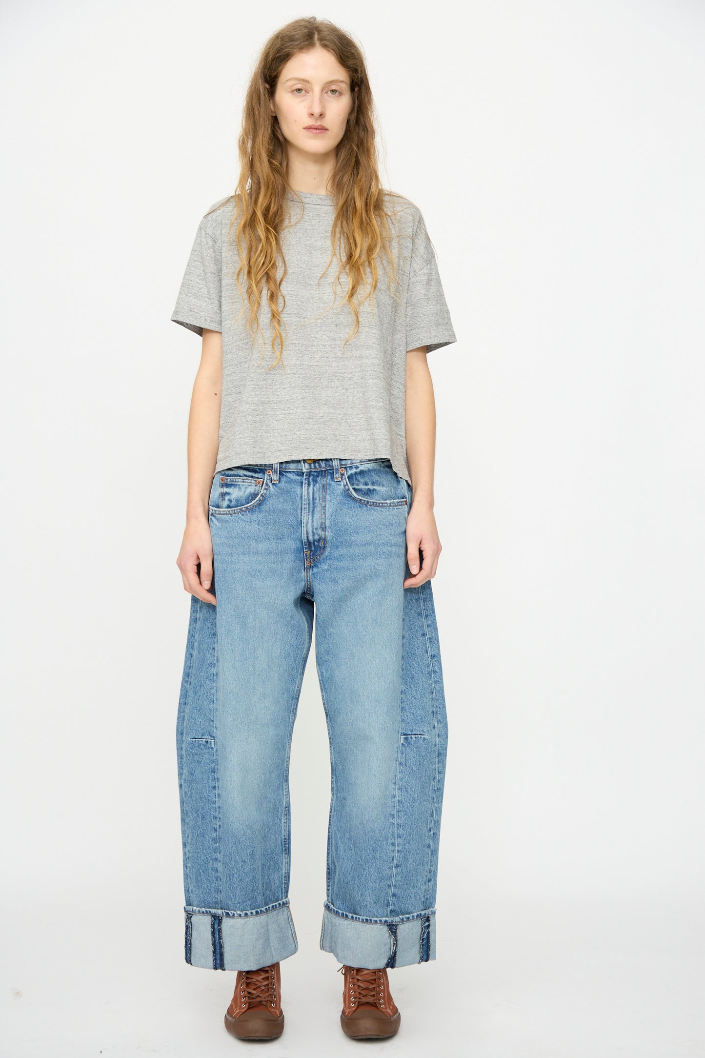 A person stands facing forward against a plain white background, wearing a gray t-shirt, B Sides Relaxed Cuffed Lasso Jean in Jane Wash (loose, wide-leg, heavy cotton denim with large cuffs), and brown lace-up boots.