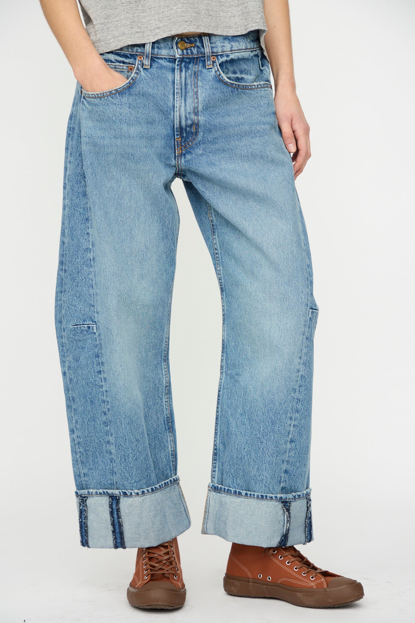 A person wears B Sides Relaxed Cuffed Lasso Jean in Jane Wash—light blue wide-leg jeans with large cuffs—paired with a gray t-shirt and brown lace-up shoes, standing against a plain background.