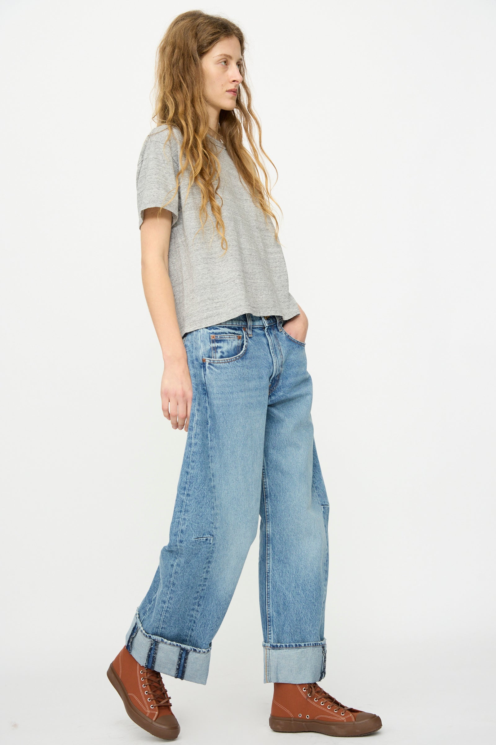 A person with long hair, in a gray T-shirt, B Sides Relaxed Cuffed Lasso Jean in Jane Wash, and brown high-top sneakers stands against a plain white background.
