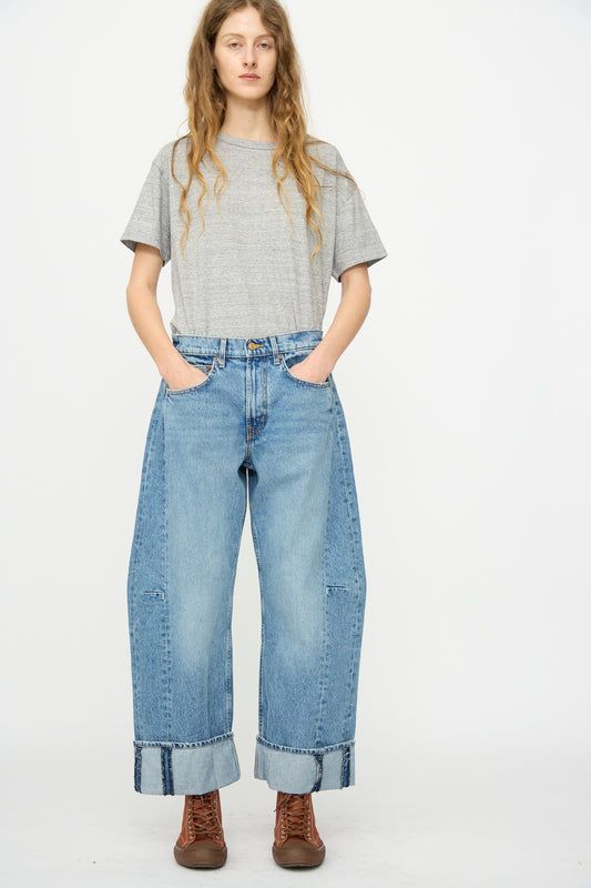 A person stands against a plain background wearing a gray t-shirt, brown boots, and B Sides’ Relaxed Cuffed Lasso Jean in Jane Wash with hands in their pockets.