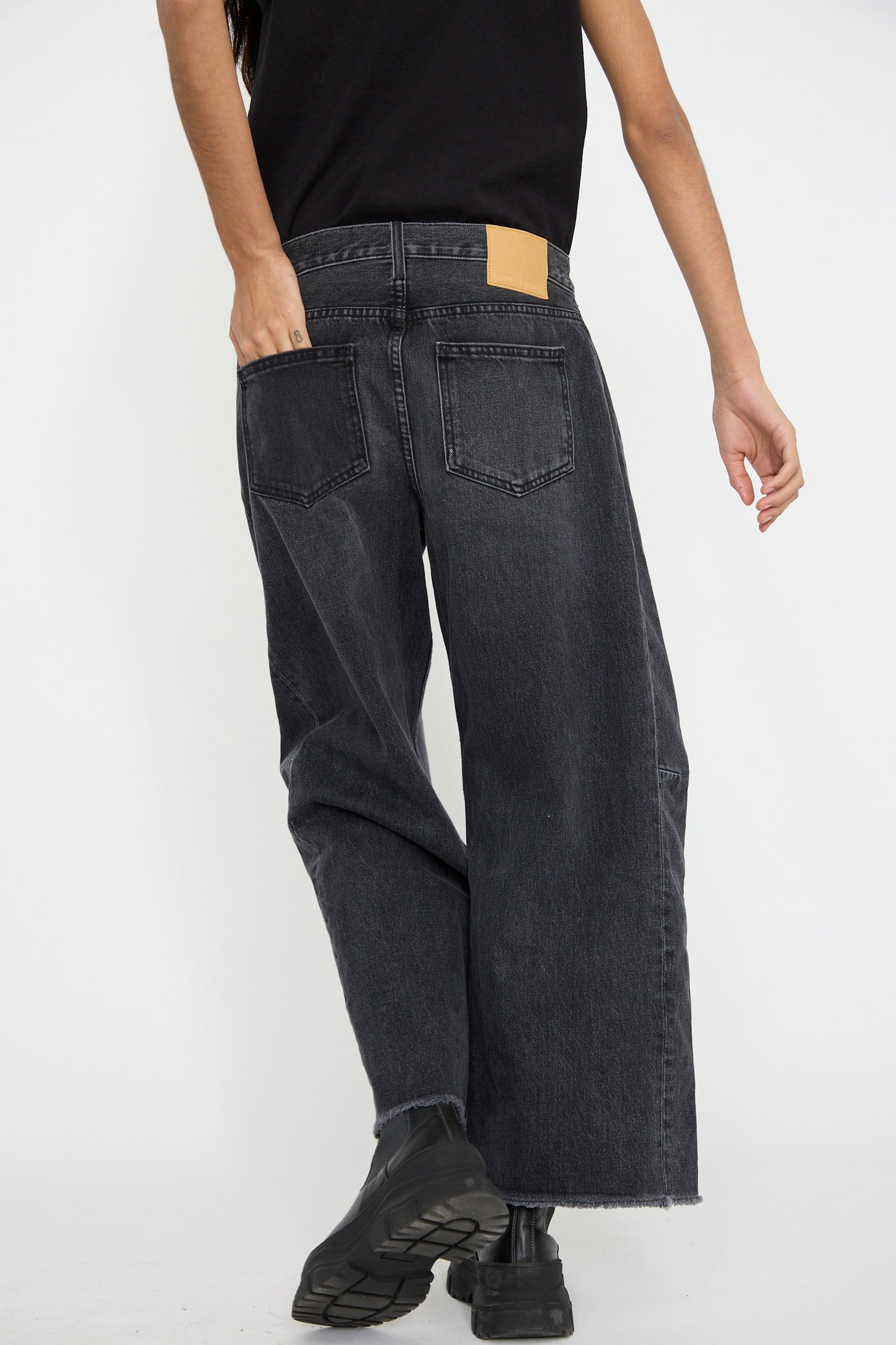 A person is wearing the Relaxed Lasso Jean in Stil Black, made from 100% cotton by B Sides, along with black boots, as viewed from behind against a white background.