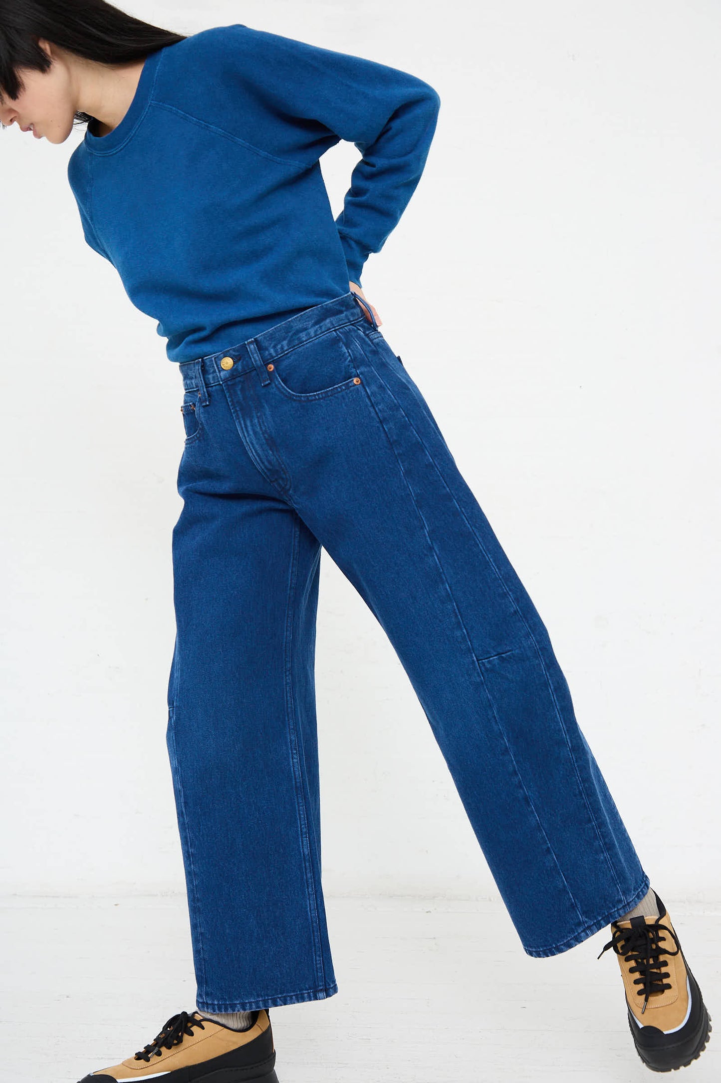 A person wearing a blue sweatshirt and the B Sides Relaxed Lasso Jean in Van Blue, posing with a bent arm, and black and tan shoes, against a white background.