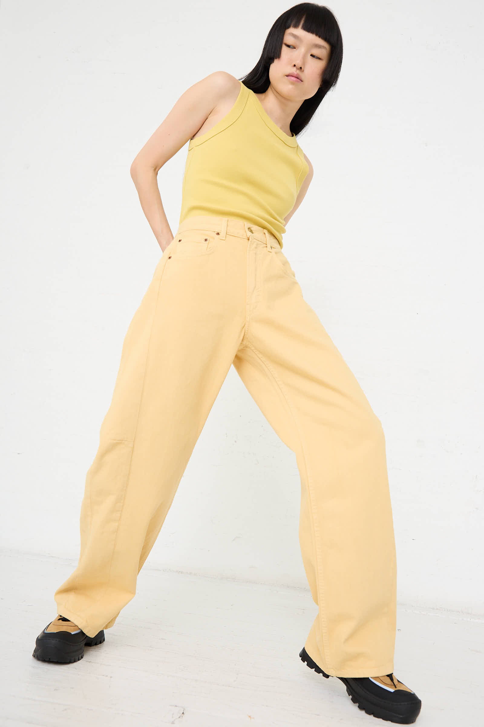 A person stands against a plain white background wearing a yellow tank top, beige wide-leg pants, and black shoes while striking a pose in the Relaxed Lasso Long Jean in Yellow Overdye by B Sides.