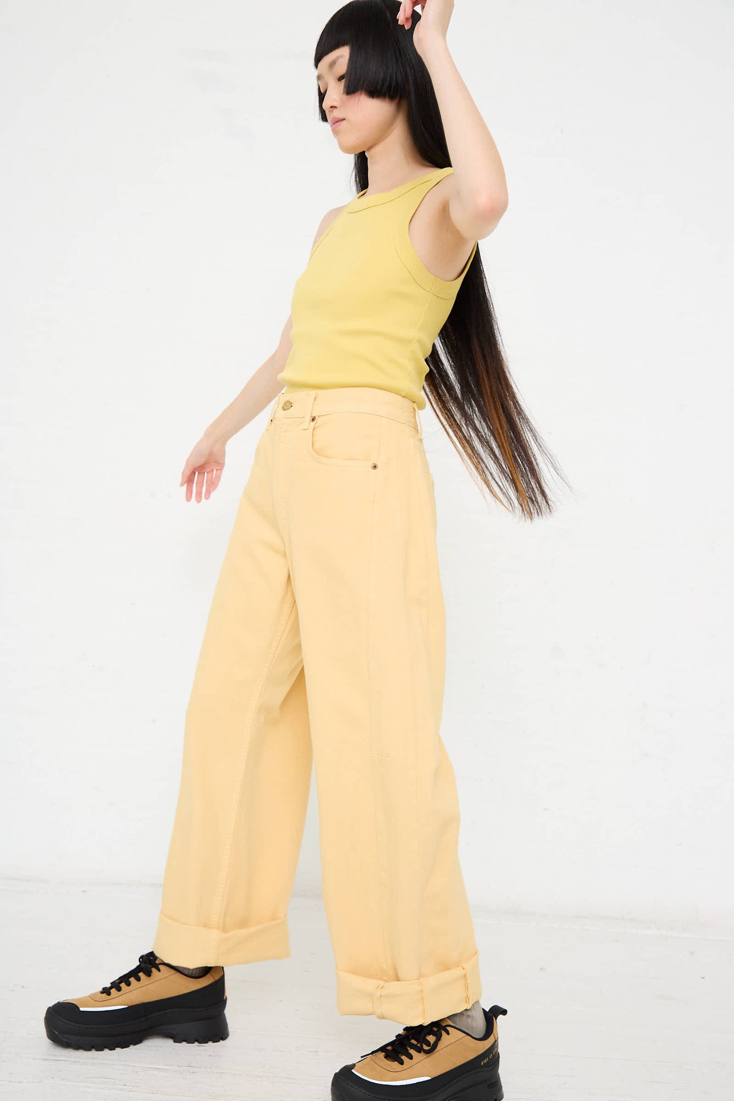 Person with long hair wearing a yellow sleeveless top, the B Sides Relaxed Lasso Long Jean in Yellow Overdye made from 100% cotton denim, and black-and-brown platform shoes, standing against a plain white background.