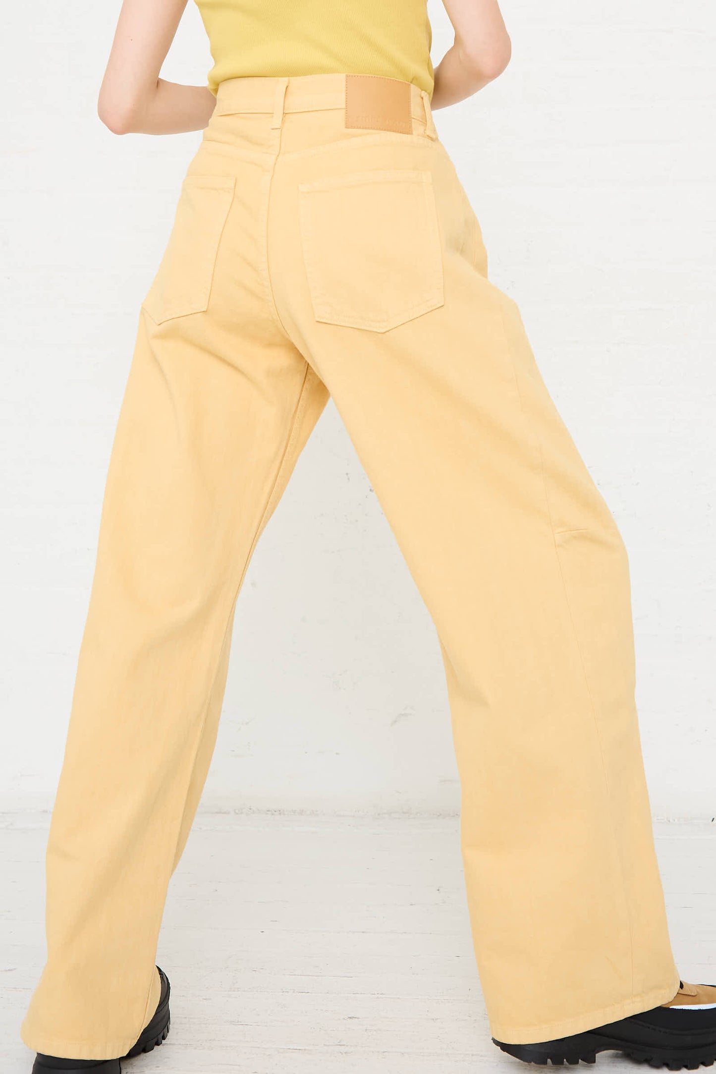 A person wearing a yellow sleeveless top and the Relaxed Lasso Long Jean in Yellow Overdye by B Sides, standing against a white background.